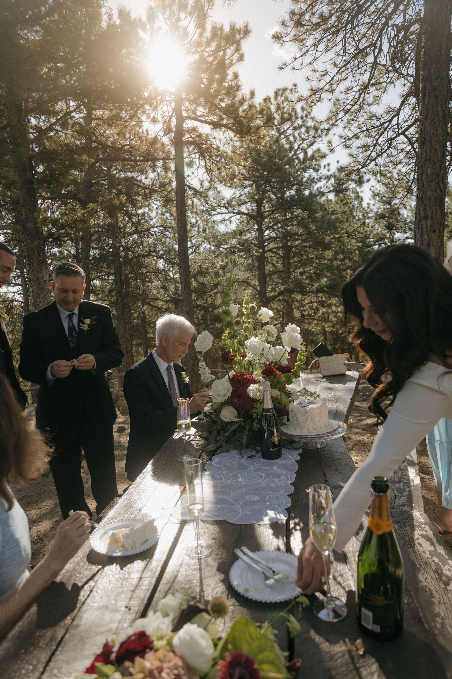 Outdoor wedding reception on a rustic wooden picnic table in a pine forest with floral centerpieces and champagne.