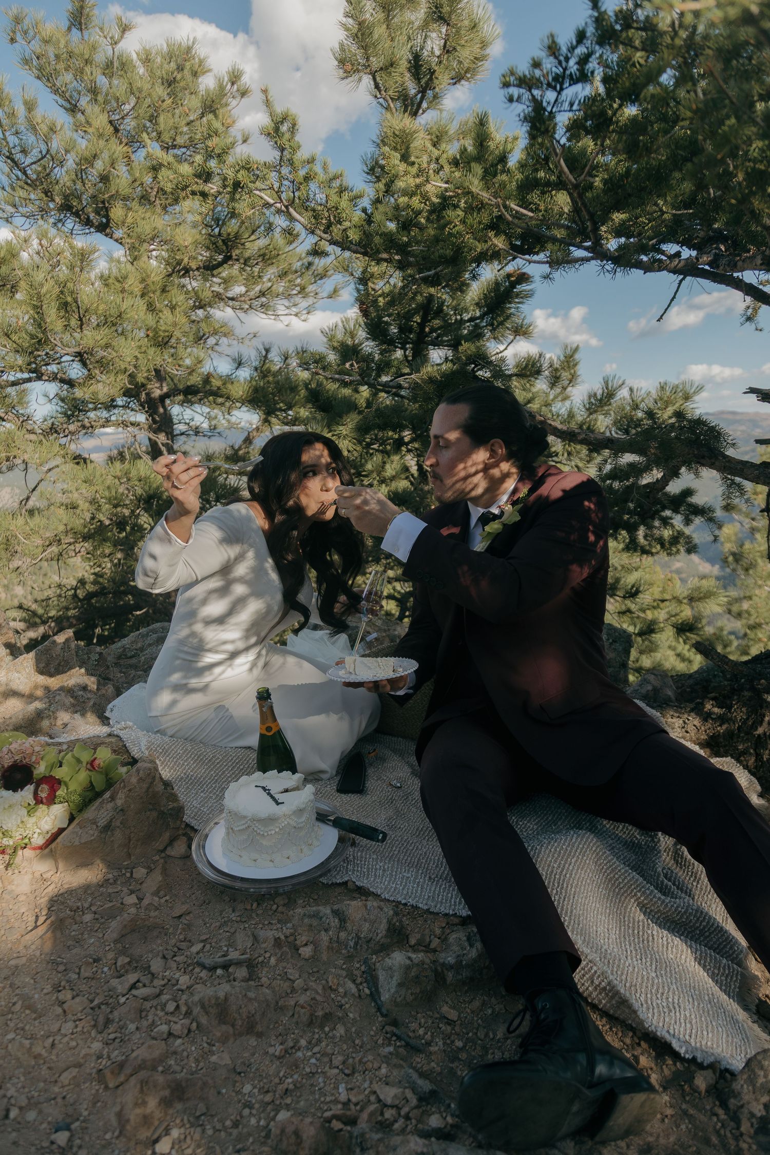 Bride and groom sharing wedding cake during an outdoor elopement picnic surrounded by pine trees.