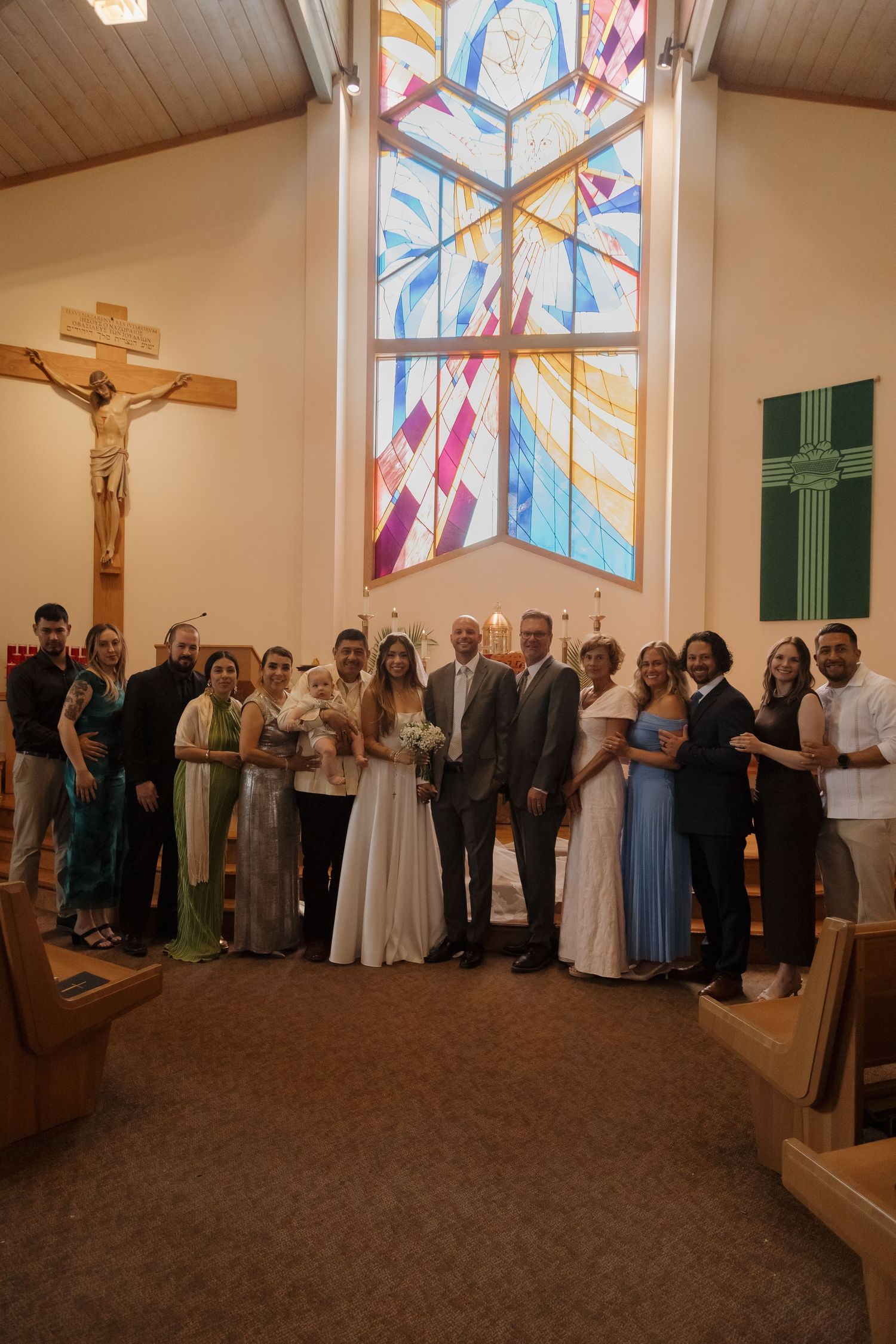 Wedding party posing together inside church with colorful stained glass windows and crucifix in background.