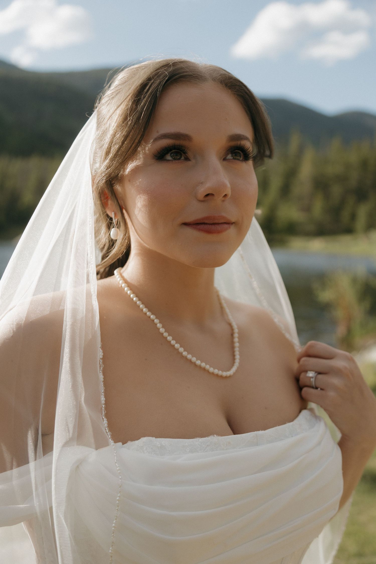 Elegant bride in white strapless gown with pearl necklace and veil poses against a scenic mountain lake backdrop.