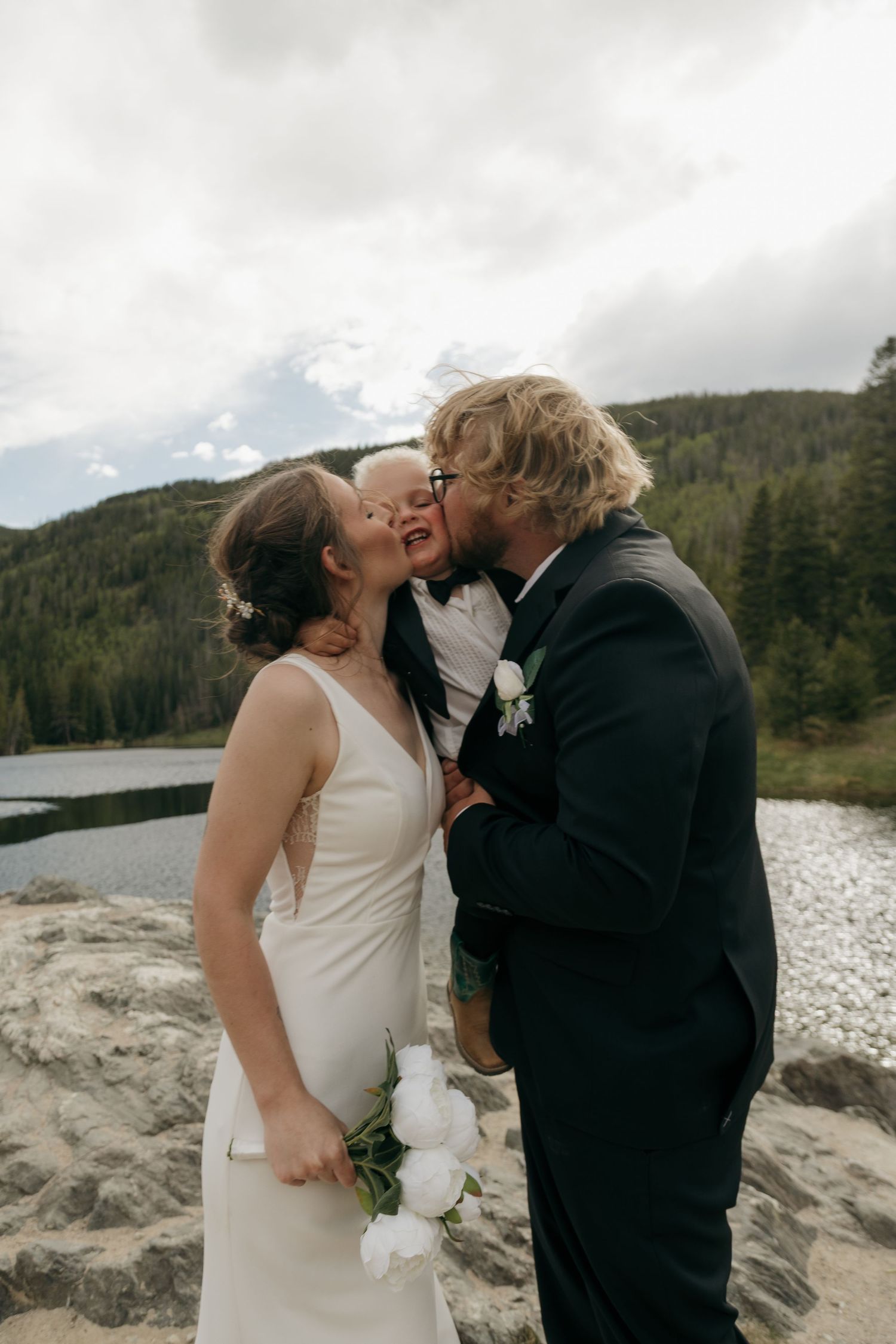 Bride and groom share joyful moment kissing young child in tuxedo by mountain lake.