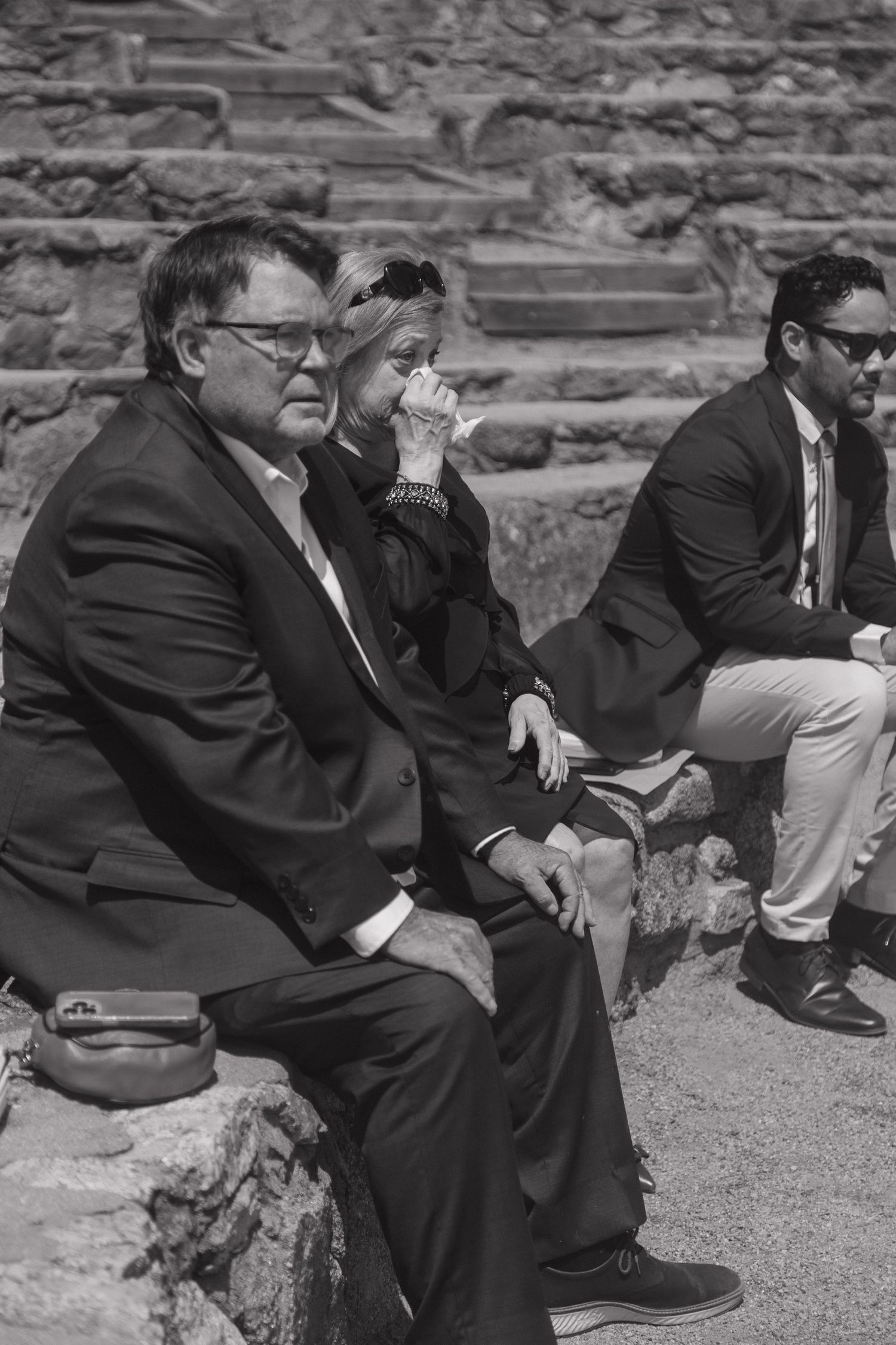Black and white photo of wedding guests seated on stone steps, with a man in a suit wiping his face with a tissue.