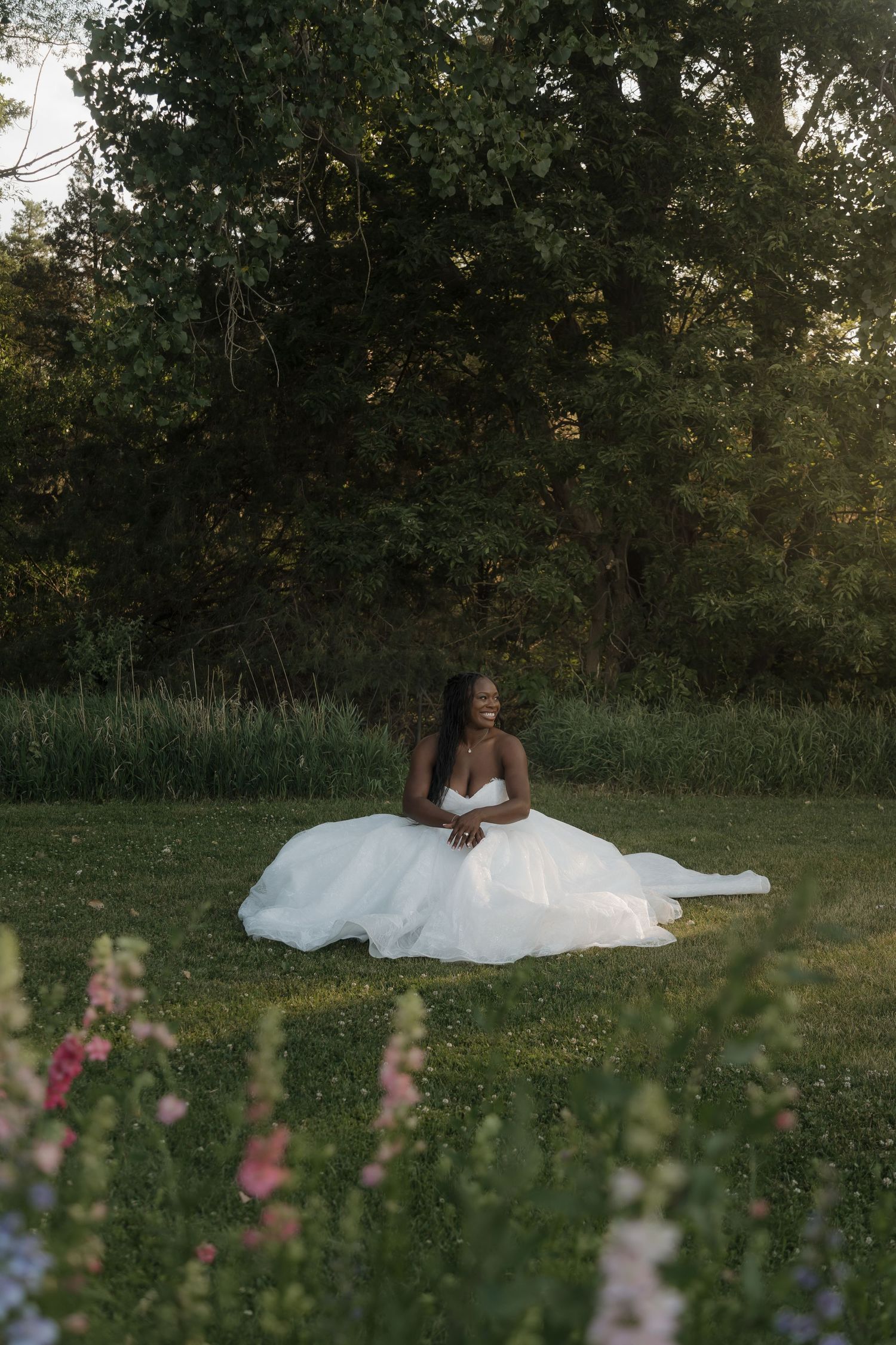 Black bride in white ballgown sits gracefully on green grass surrounded by pink flowers and lush trees at golden hour.