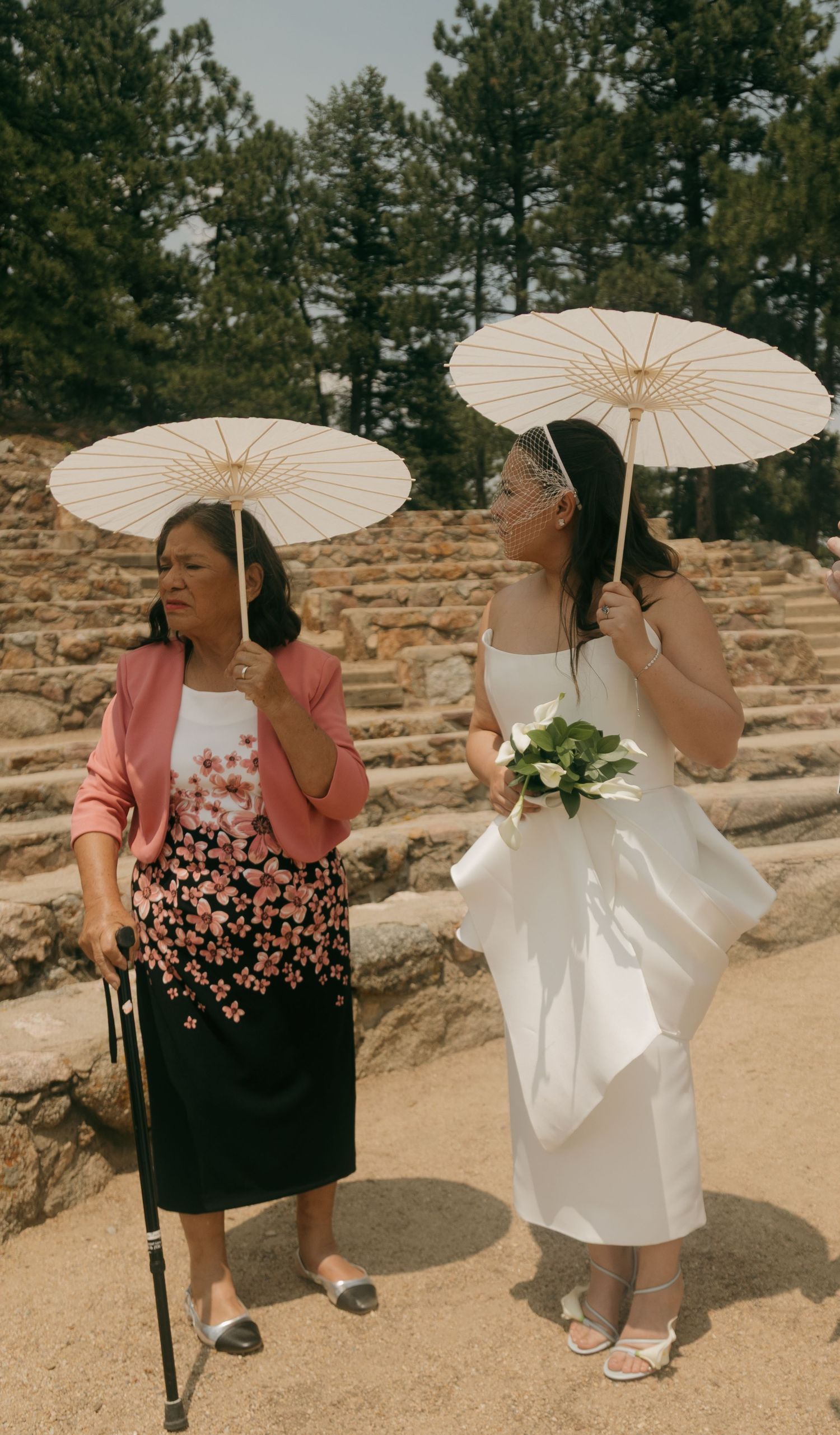 Bride in white dress holding bouquet and parasol walks with older woman using cane near stone steps outdoors.