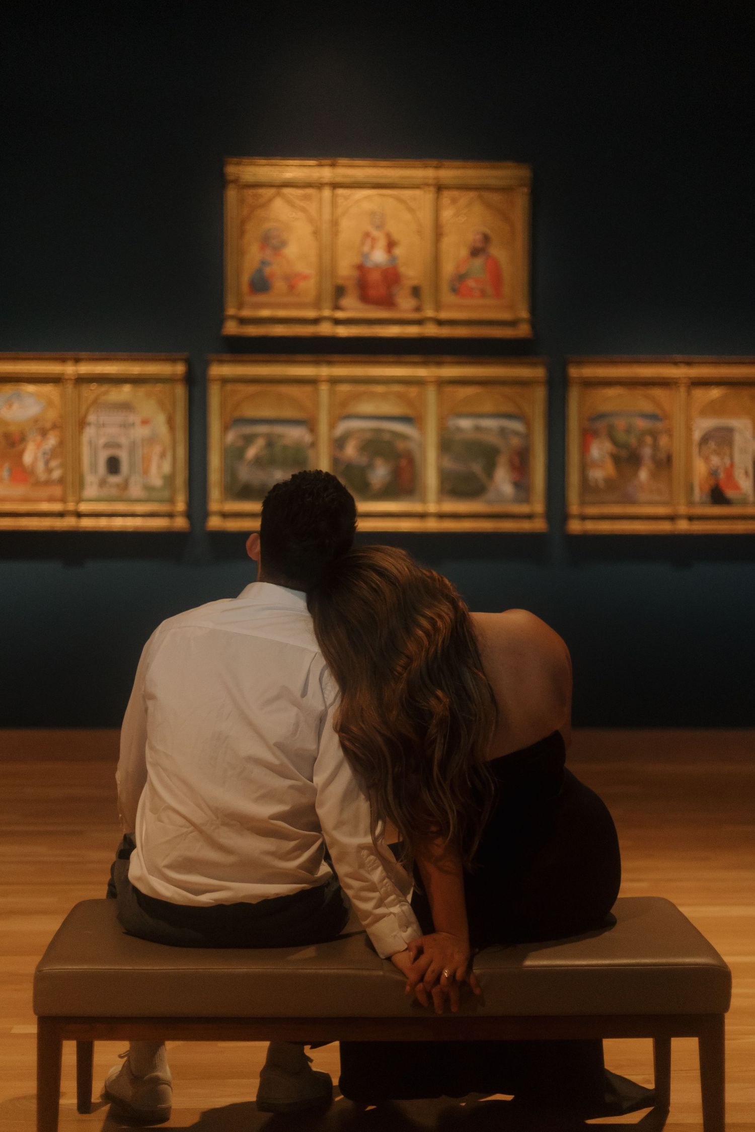 Couple sitting together on a museum bench viewing illuminated Renaissance altarpiece paintings in a dimly lit gallery.