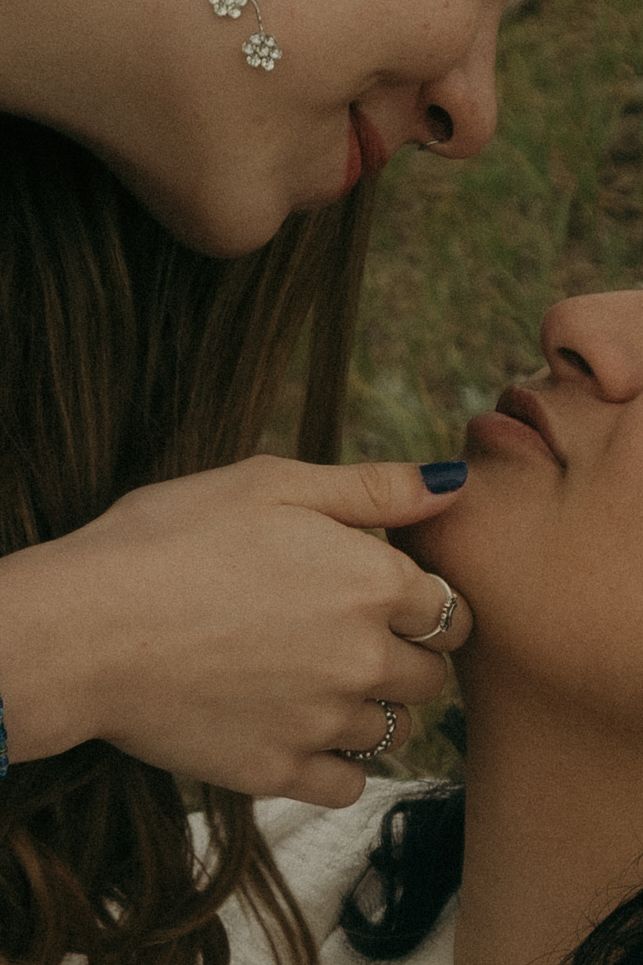 Two women sharing an intimate moment, one touching the other's chin, wearing silver rings and blue nail polish outdoors.