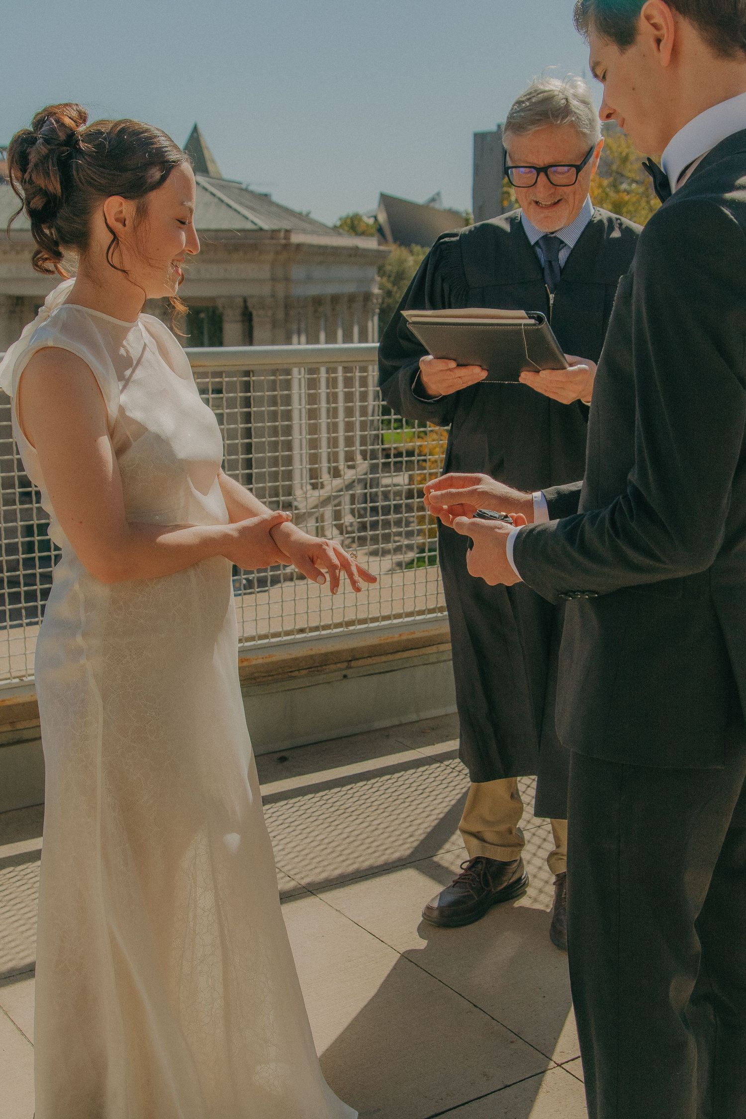 An outdoor wedding ceremony taking place on a sunny terrace with urban architecture in the background.