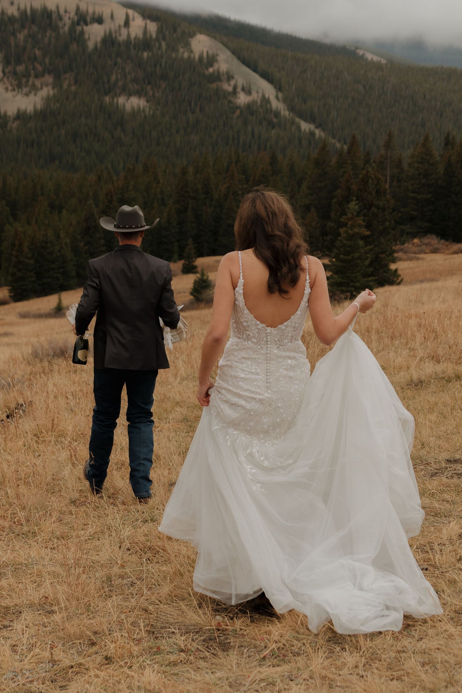 A couple walks through a mountain meadow with evergreen forests in the background.