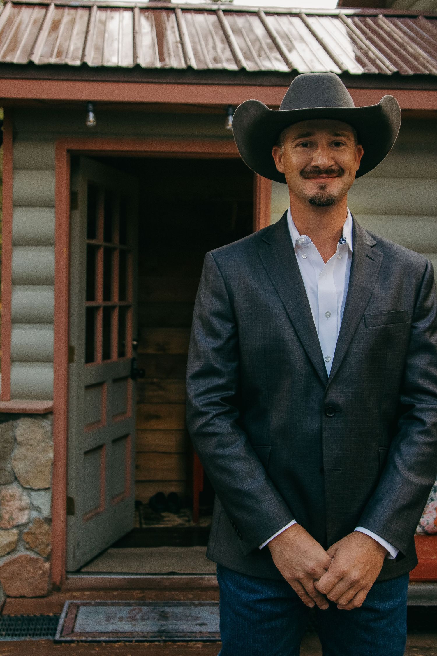 A person in a black suit and cowboy hat stands against a rustic wooden building backdrop.