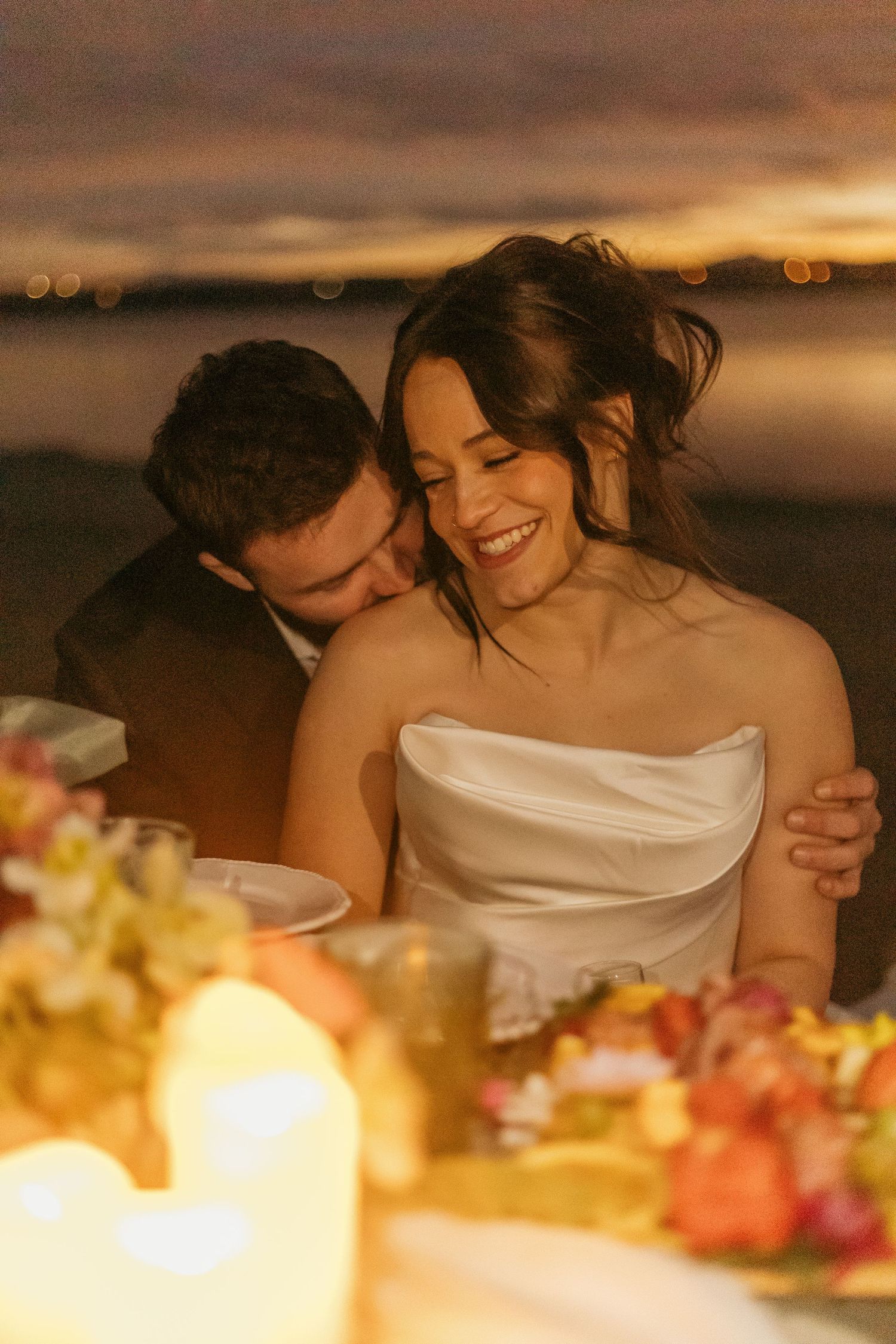 A romantic wedding couple shares an intimate moment at sunset with floral decorations in the foreground.