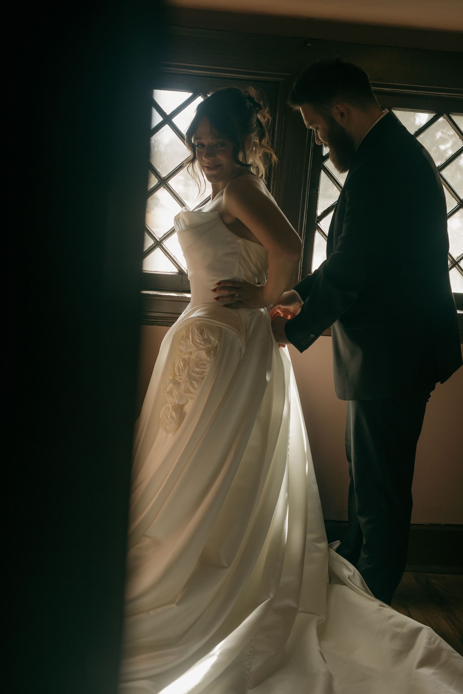 A bride in a white gown being helped with final preparations near a window.
