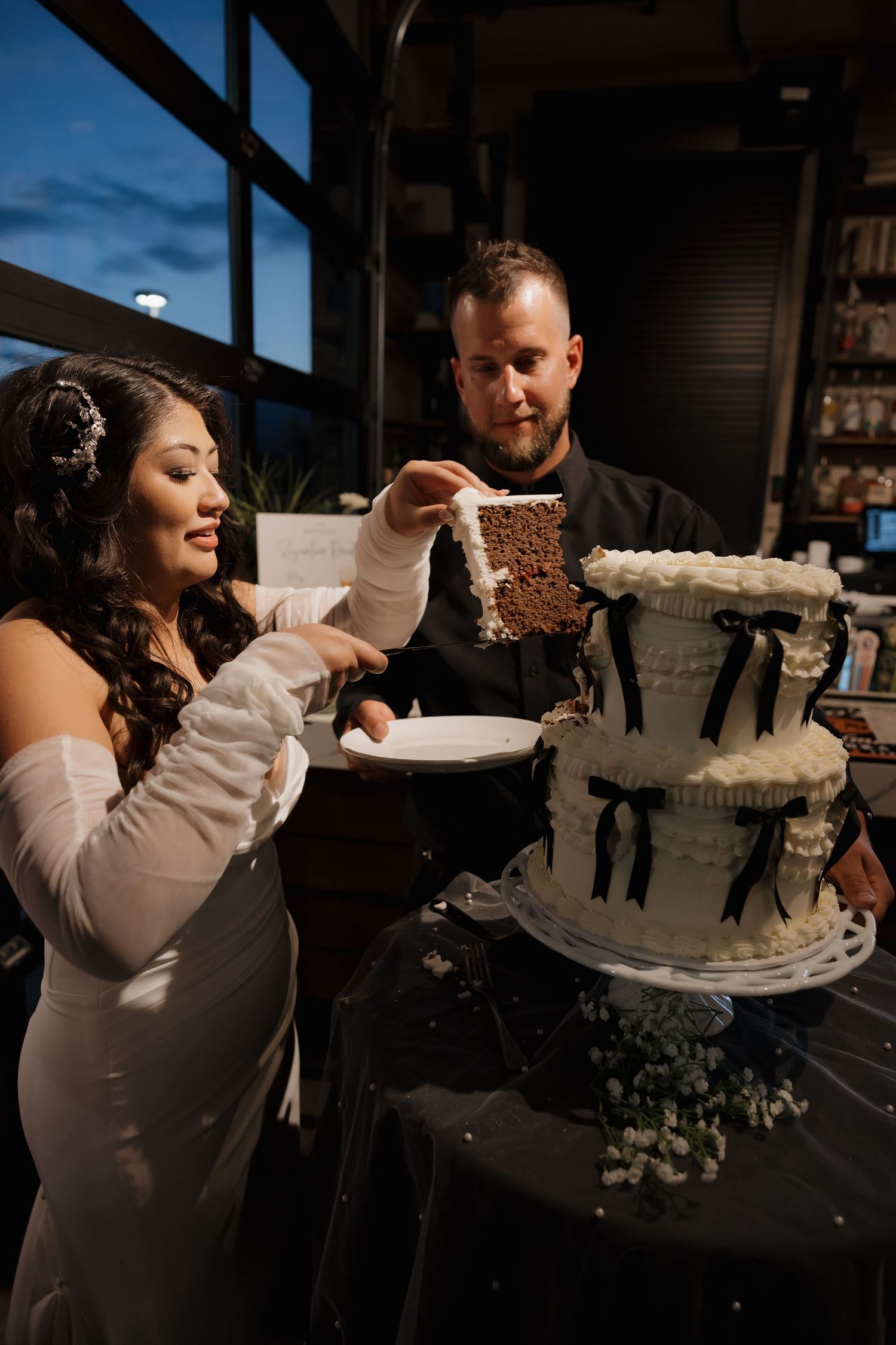 Bride and groom cut elegant three-tier white wedding cake decorated with black bows at reception.