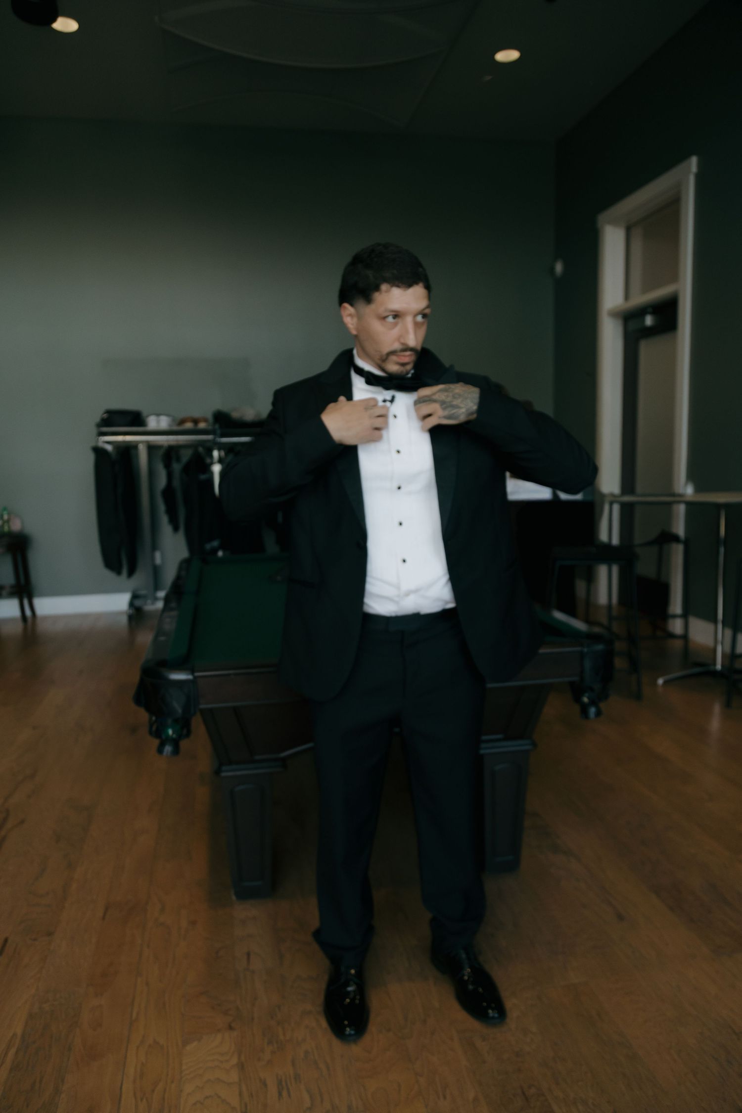 Groom in a black tuxedo adjusting his bow tie while standing in a stylish green-walled room.