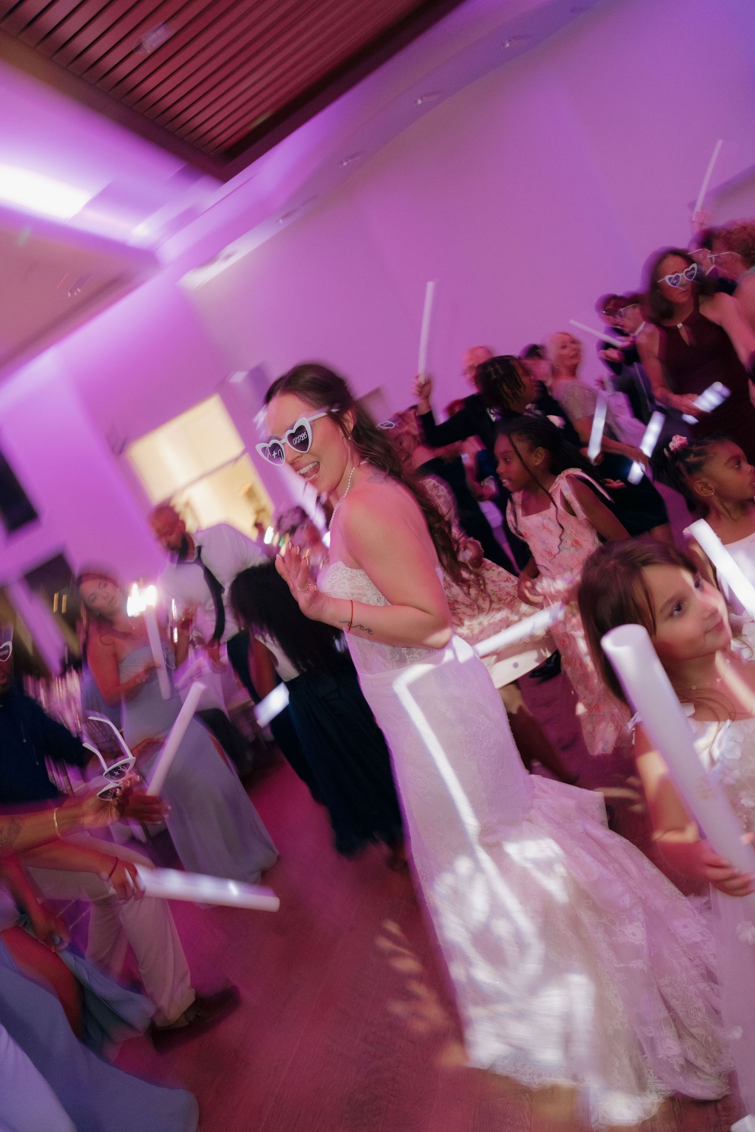 Bride in heart sunglasses dances with guests holding glow sticks under purple lights at reception.