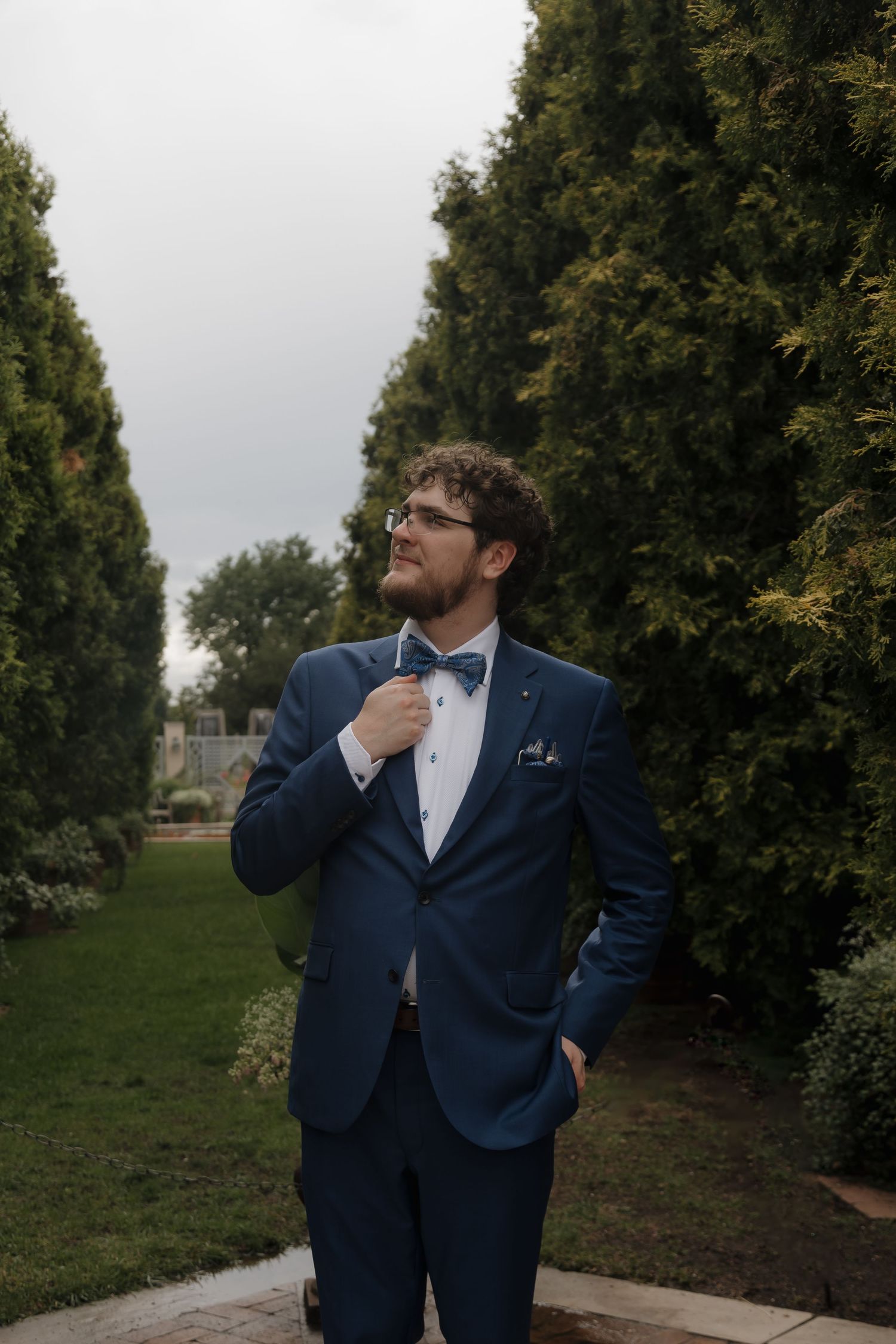 Groom in navy blue suit adjusting paisley bow tie outdoors among tall green cypress trees.