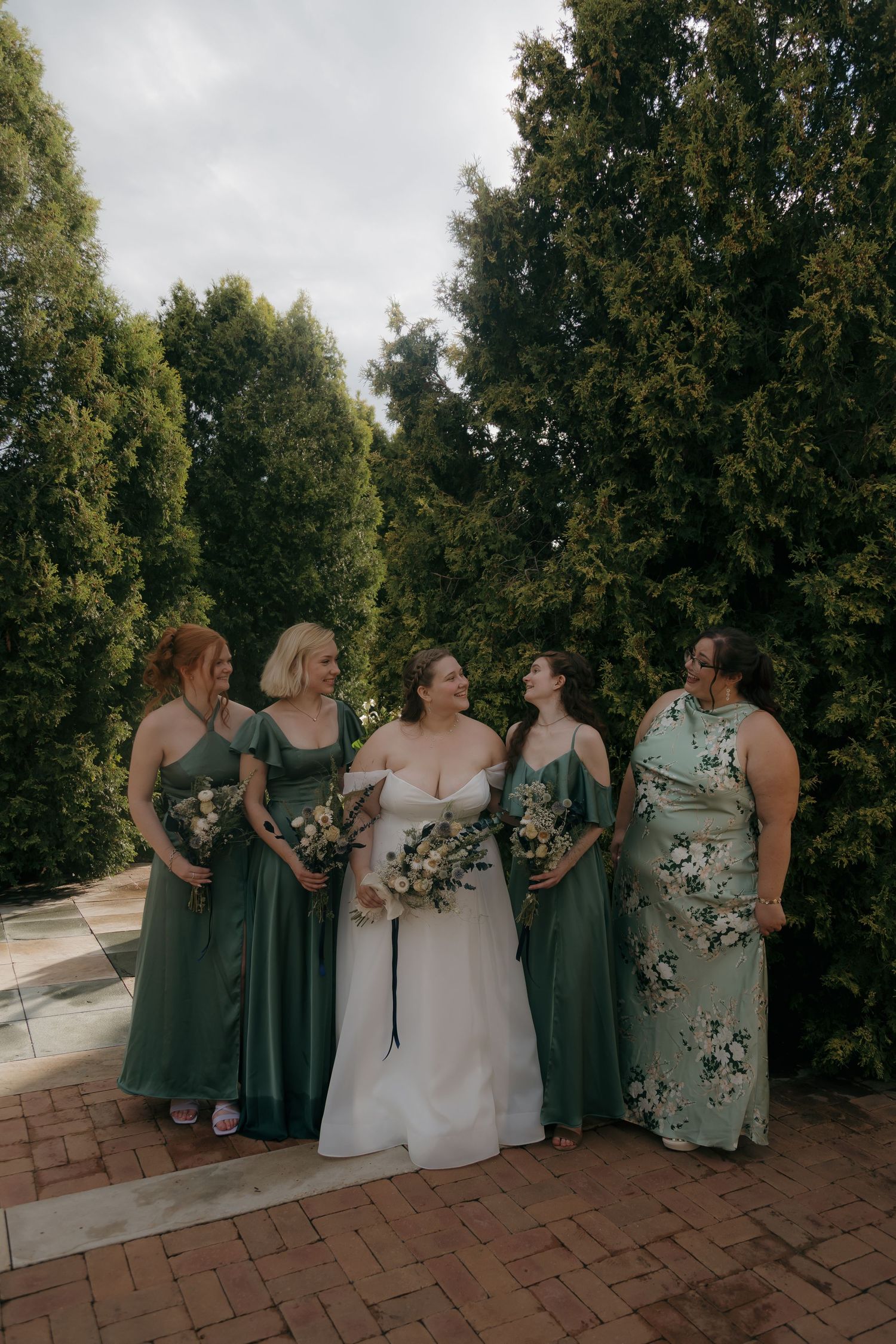 Bride in white gown poses with four bridesmaids in green dresses holding bouquets outdoors near tall evergreen trees.