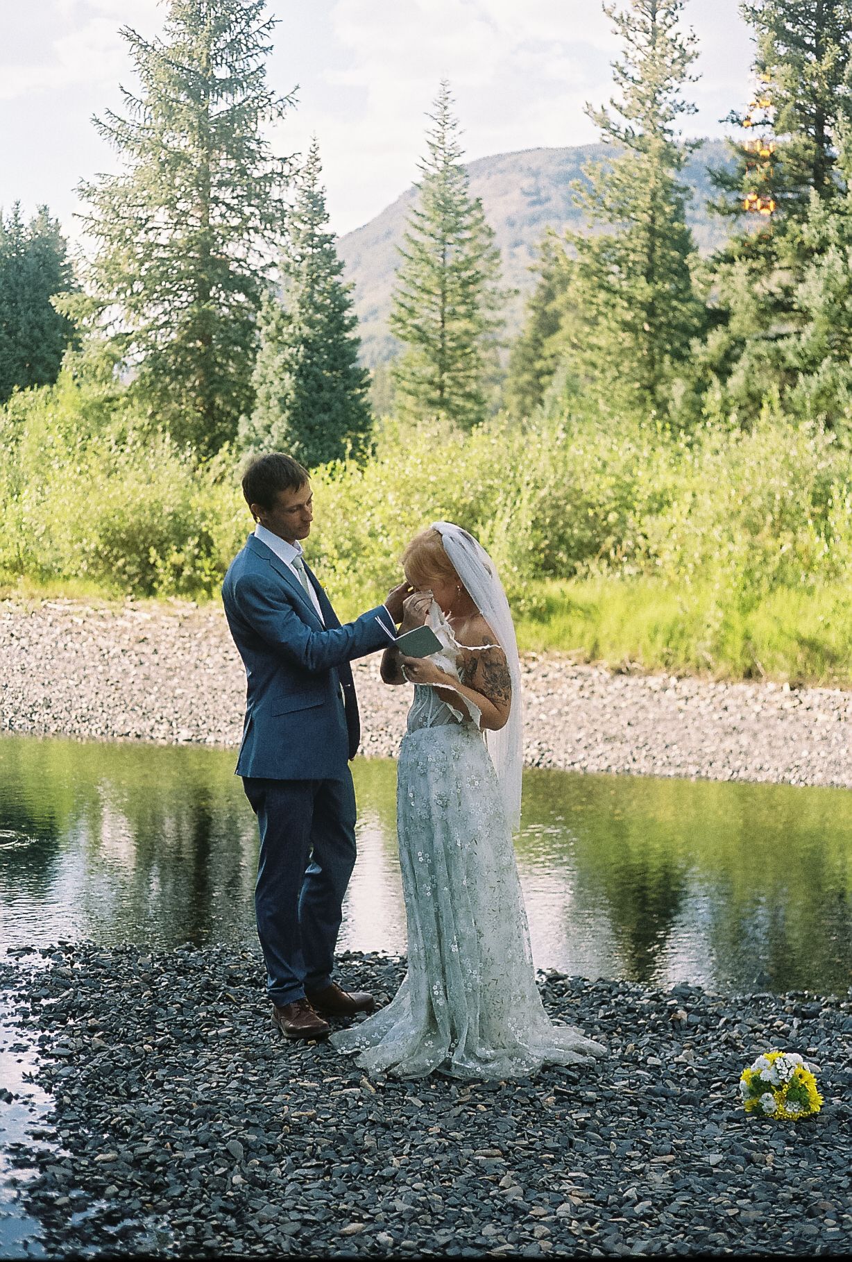 Groom places ring on bride's finger during intimate outdoor ceremony by a mountain stream in summer.
