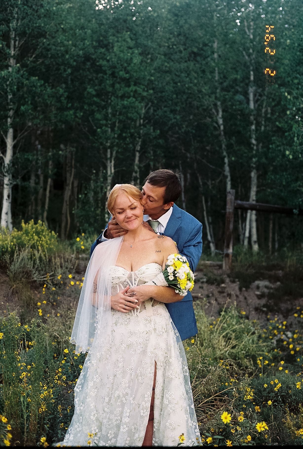 Groom kisses bride on cheek while embracing her in a wildflower meadow with aspen trees.