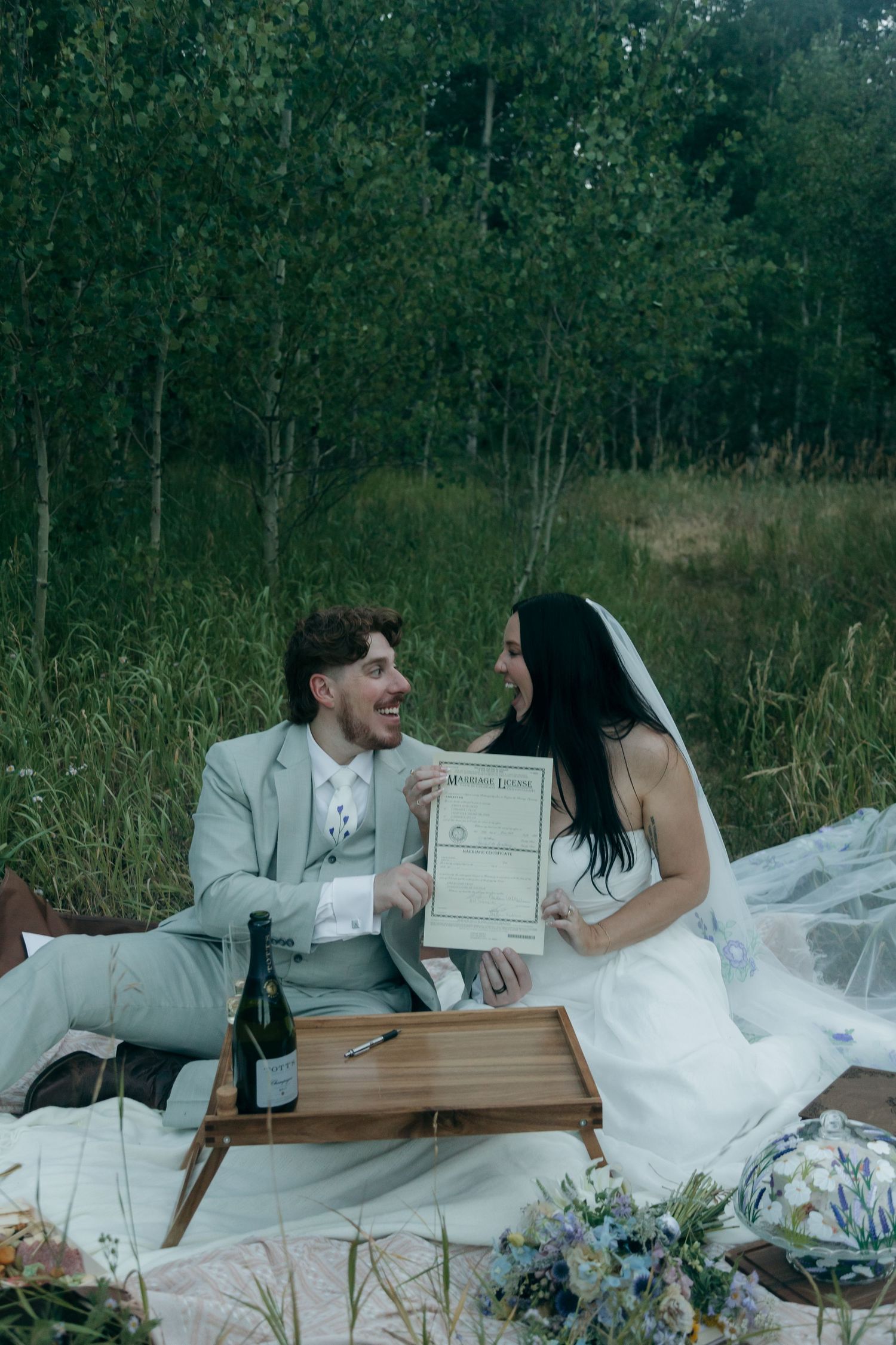 Smiling newlyweds sit on a blanket outdoors holding their marriage license with champagne and flowers nearby.