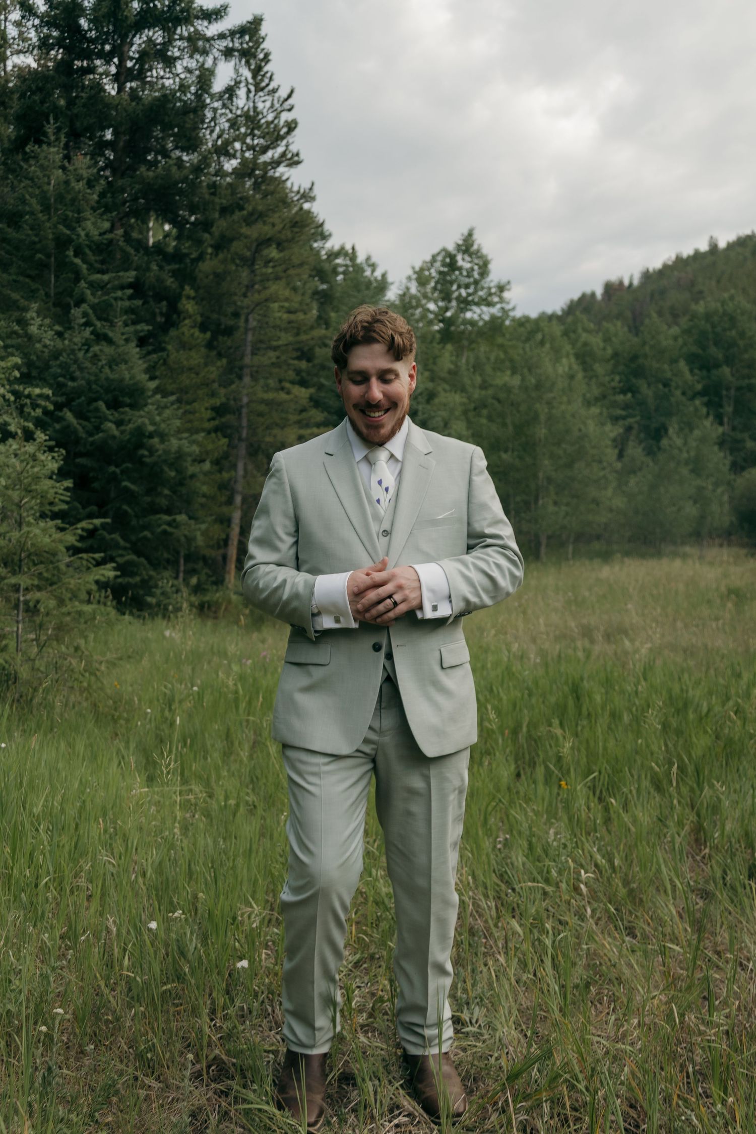 Smiling groom in sage green suit standing in lush forest meadow on cloudy wedding day.