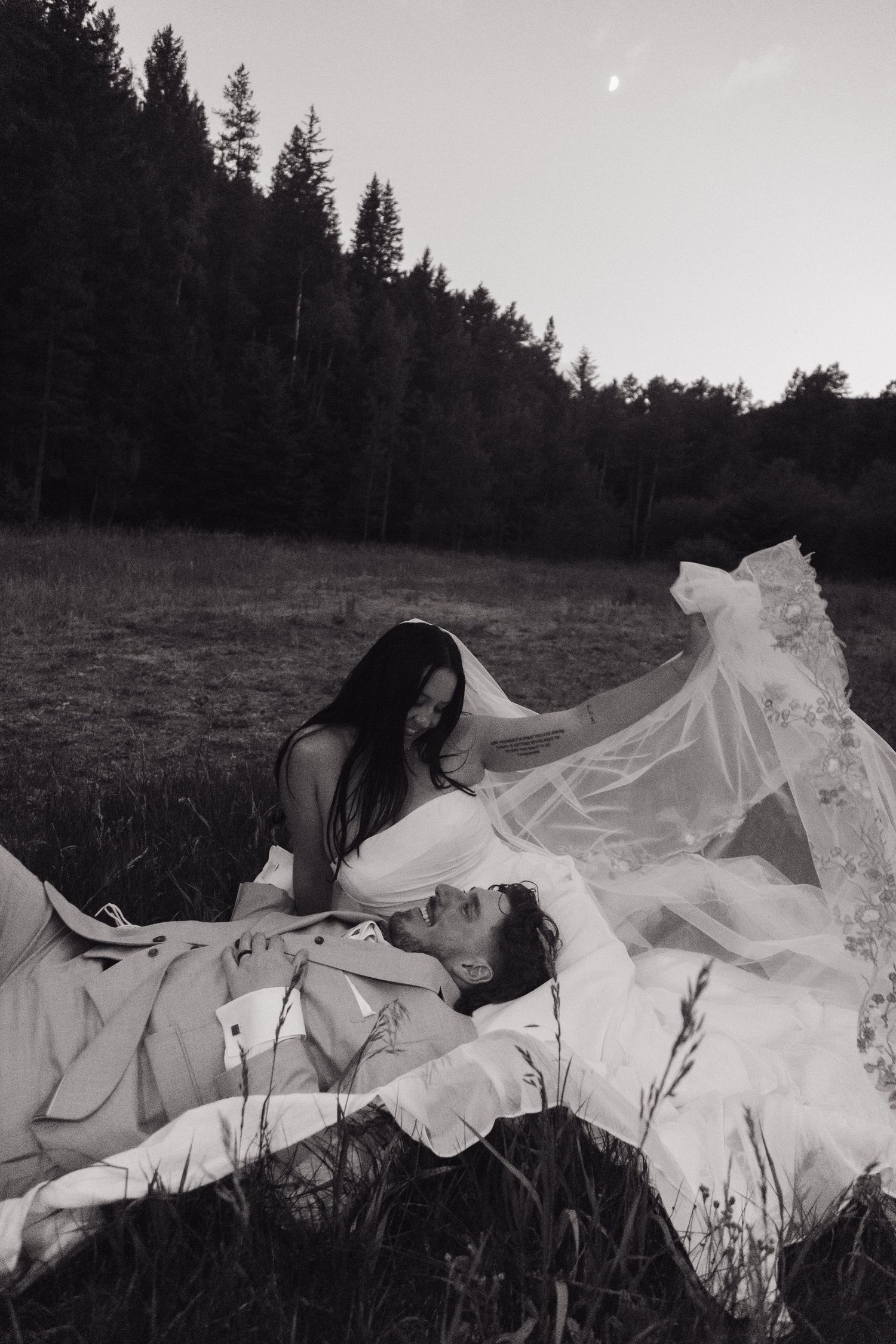 Black and white photo of bride leaning over groom lying in grass under flowing veil.