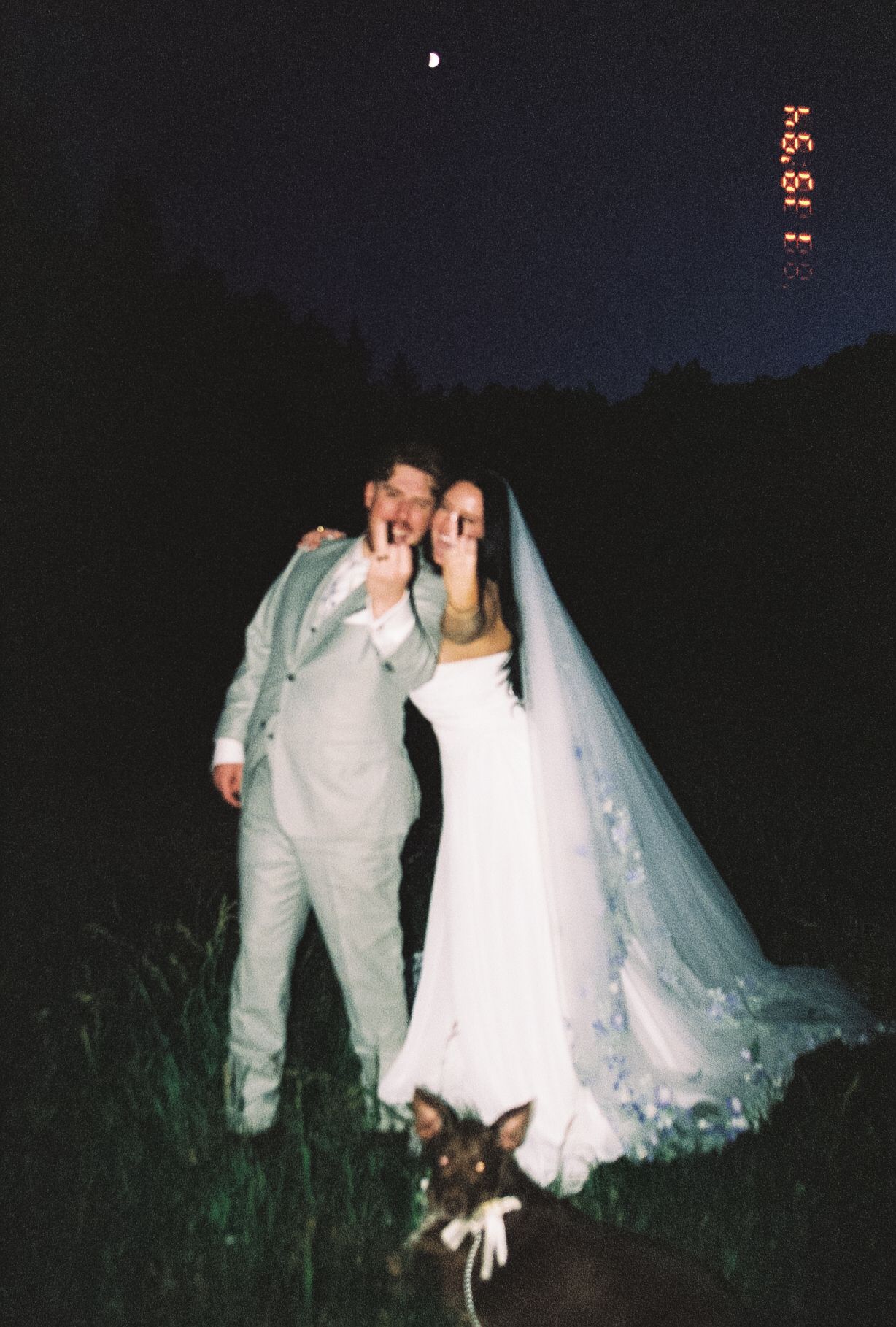 Bride and groom posing at night under moonlight with a dog at their feet in a film photography style.