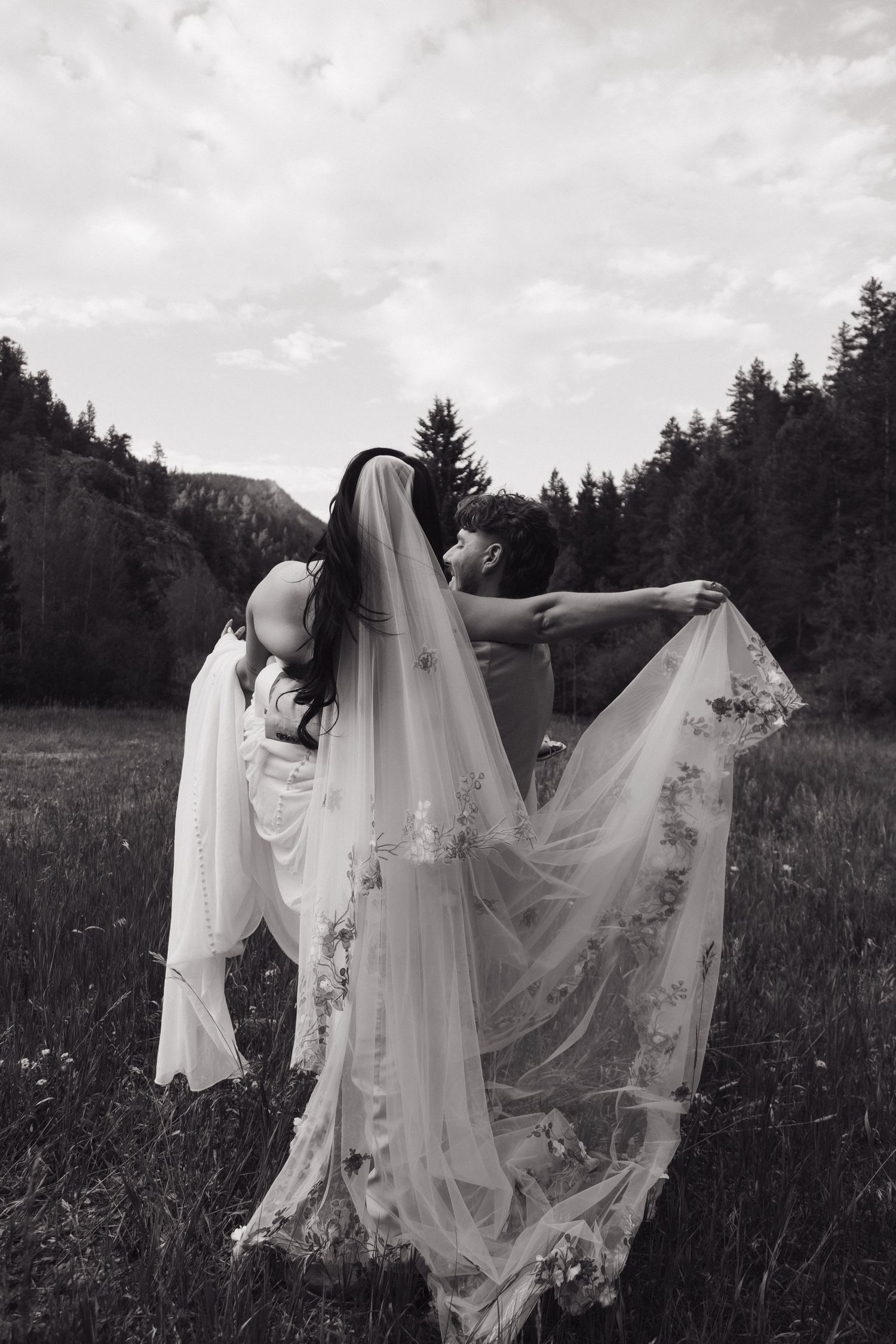 Black and white photo of bride and groom embracing in a meadow, floral veil flowing in the wind.