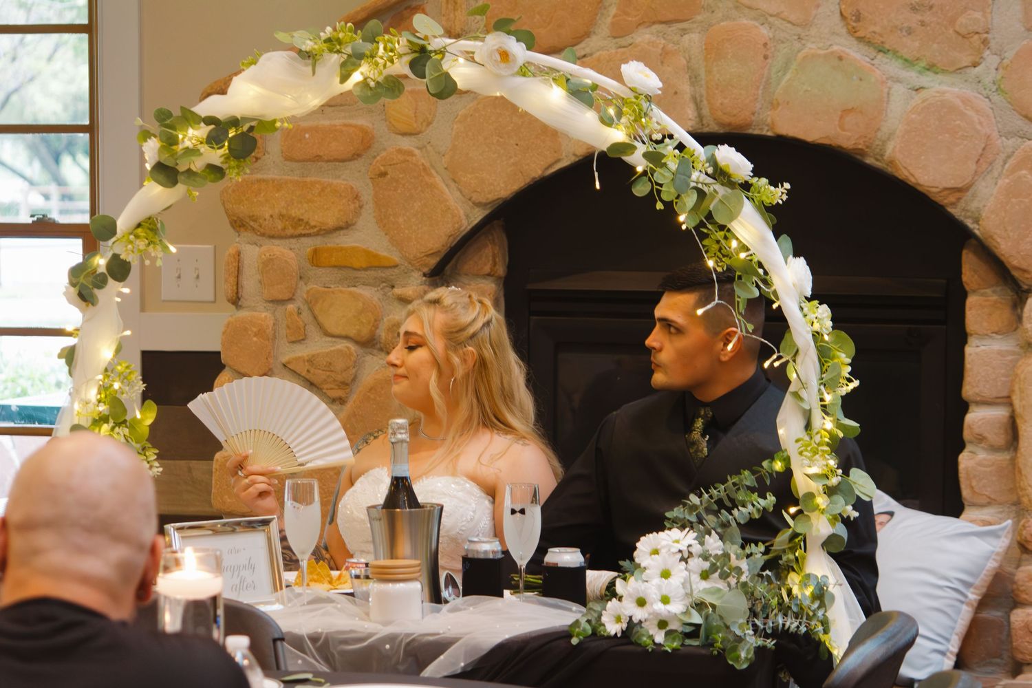 Wedding couple seated at decorated table with floral arch and stone fireplace in background.