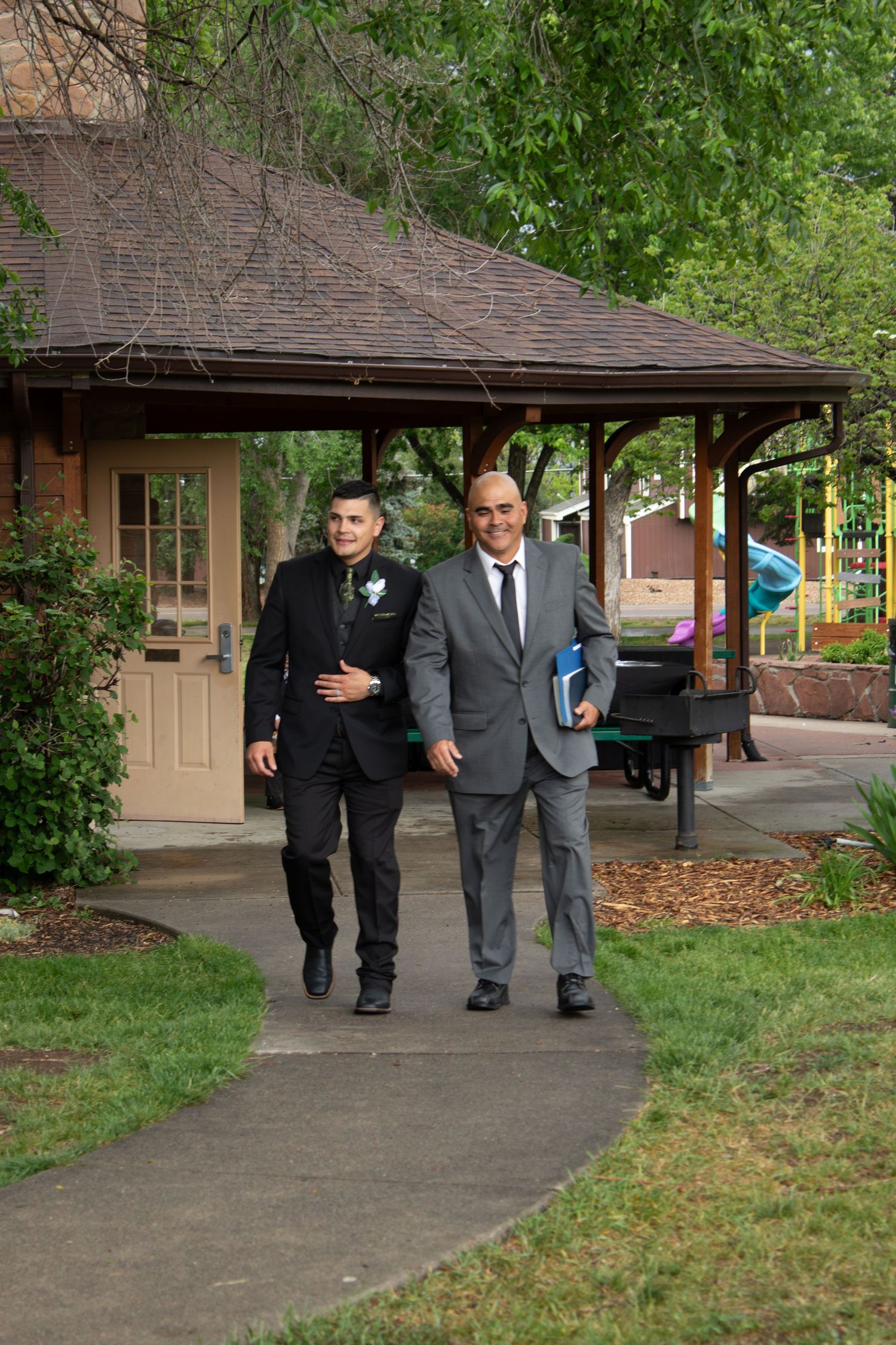 Wedding party members in formal attire walk along a covered concrete pathway outside a tan building.