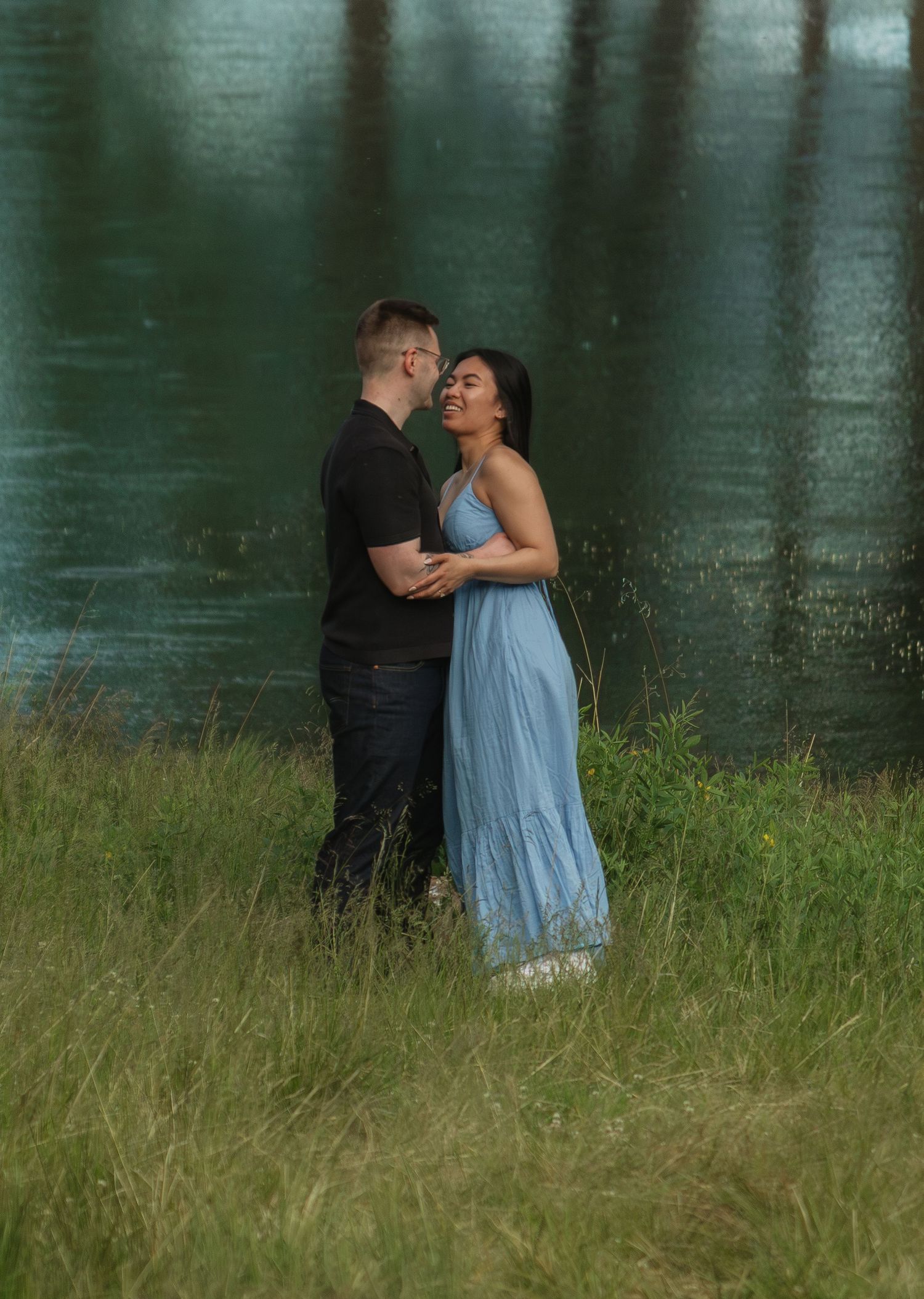 Couple sharing a tender moment by a serene lake surrounded by tall grass.