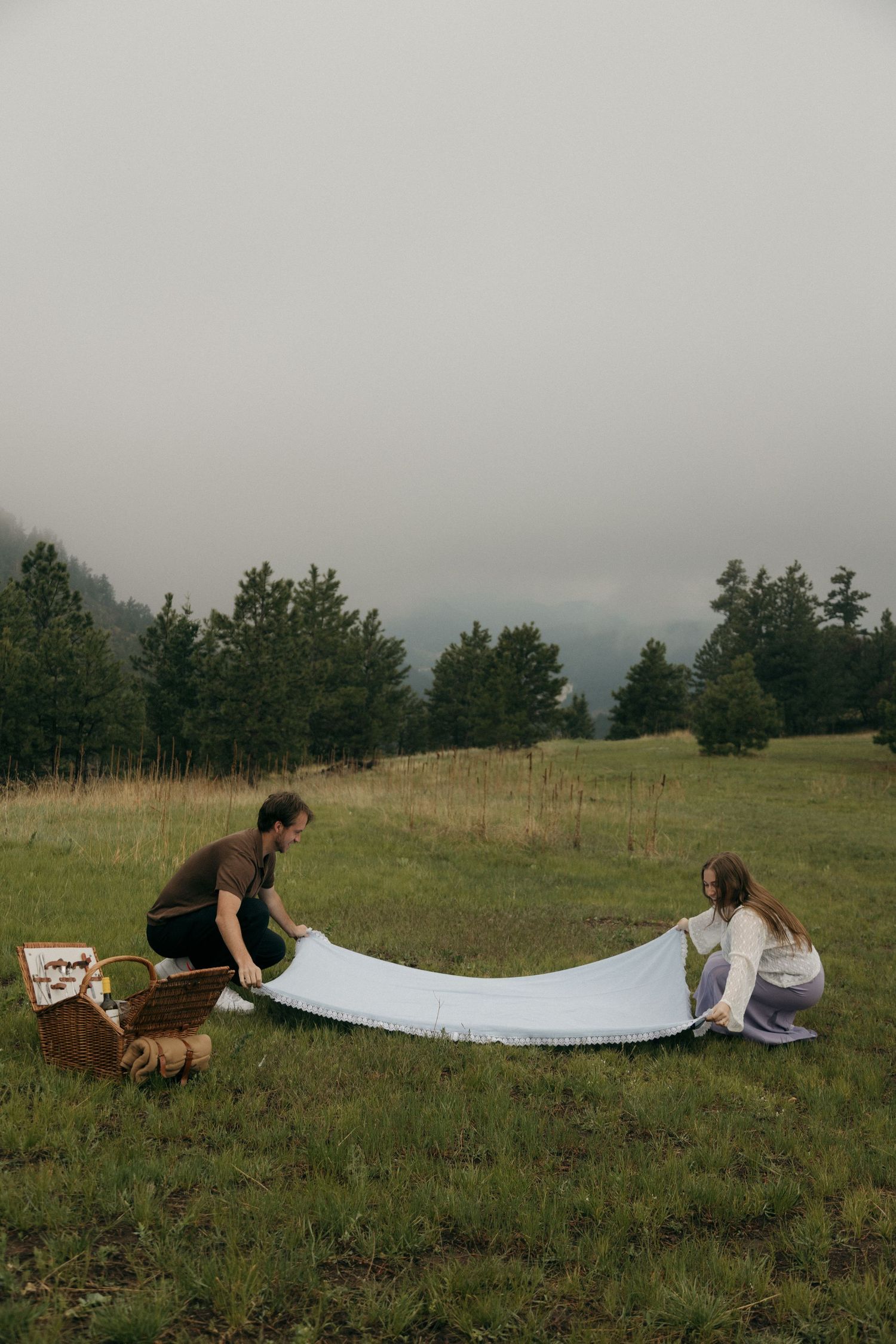 Couple spreading a white blanket on a green meadow for a picnic with a wicker basket nearby.