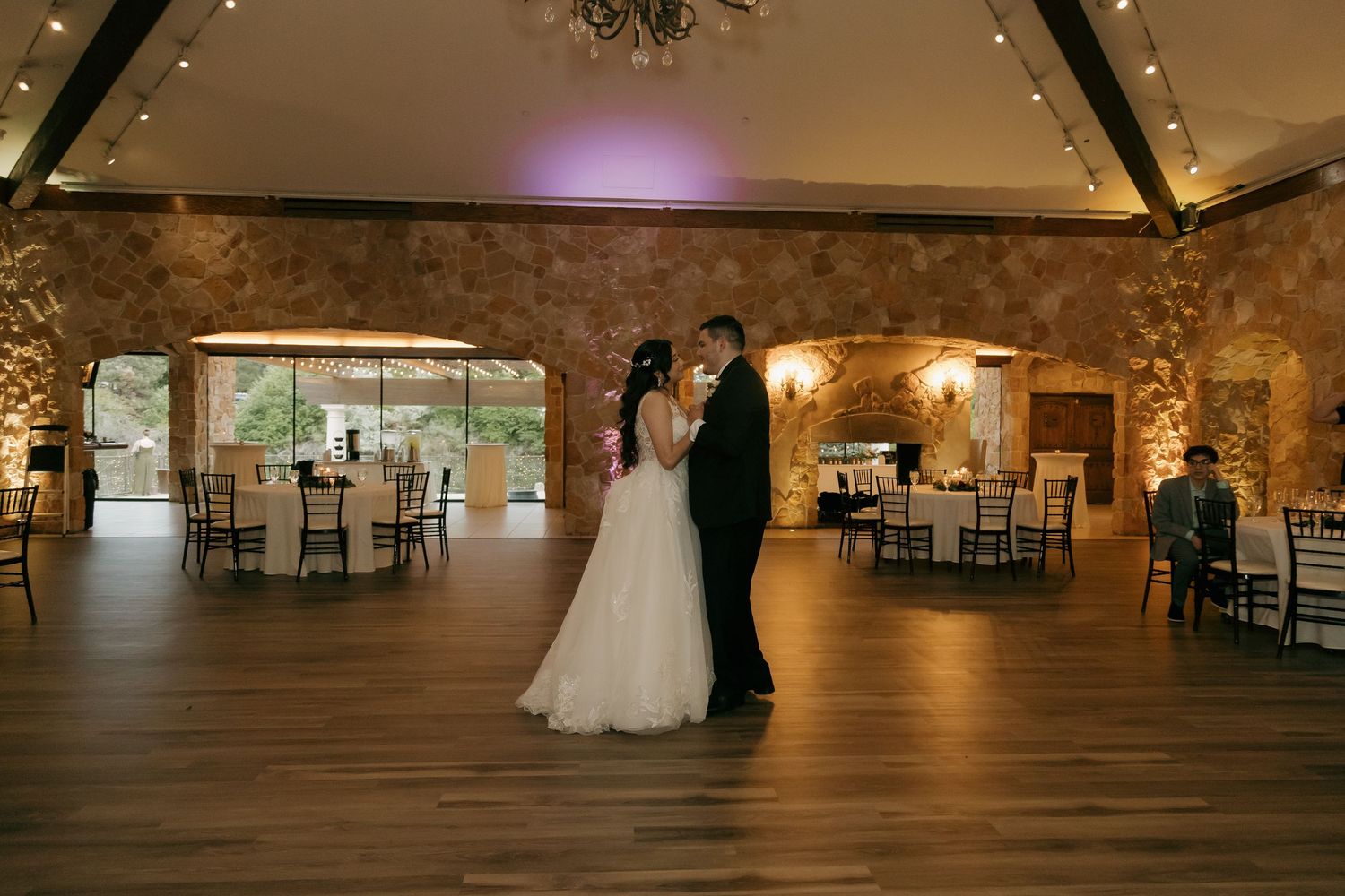 Newlyweds share intimate first dance in elegant stone-walled ballroom with warm lighting and chandelier.