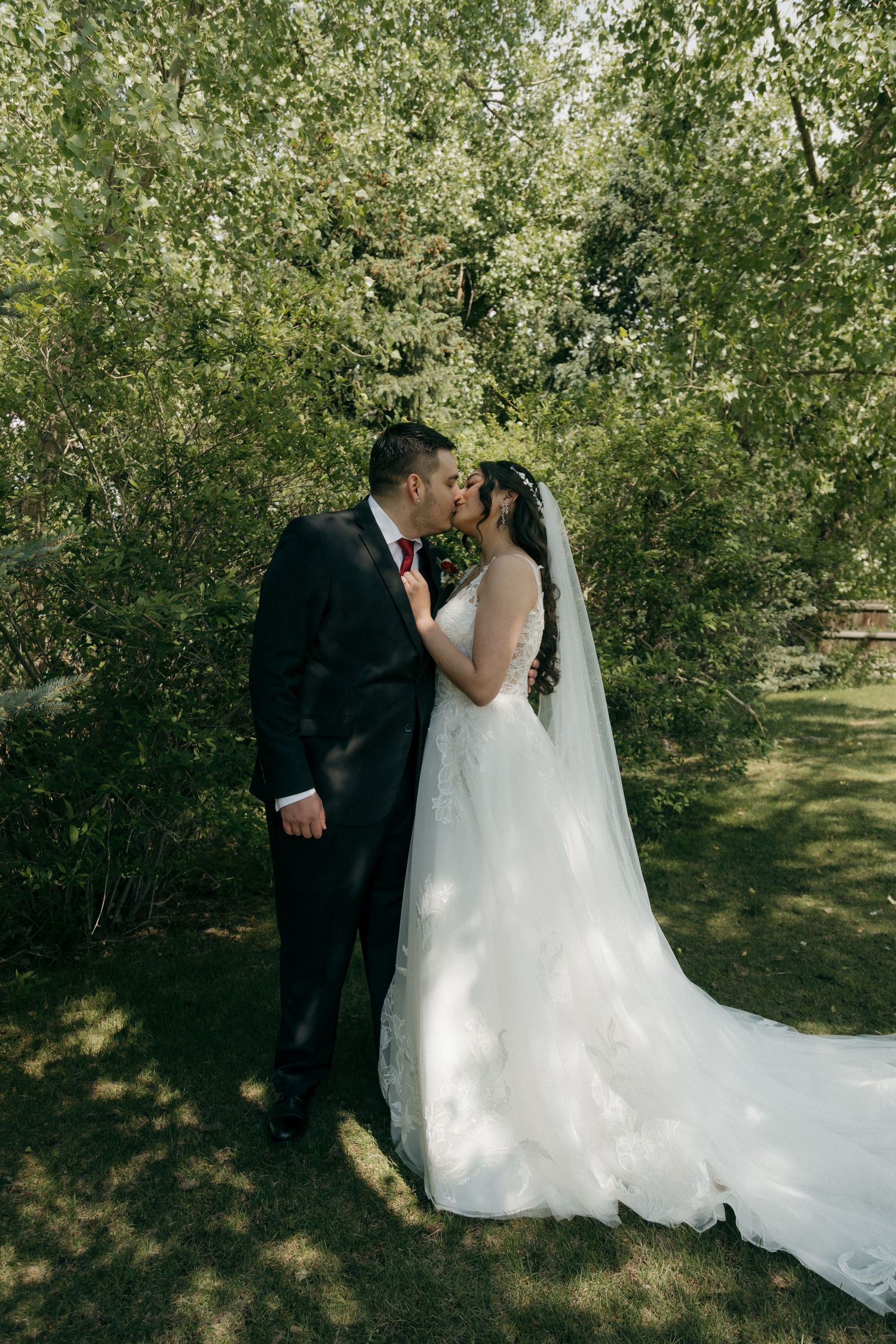 Bride and groom sharing a romantic kiss outdoors surrounded by lush green trees.