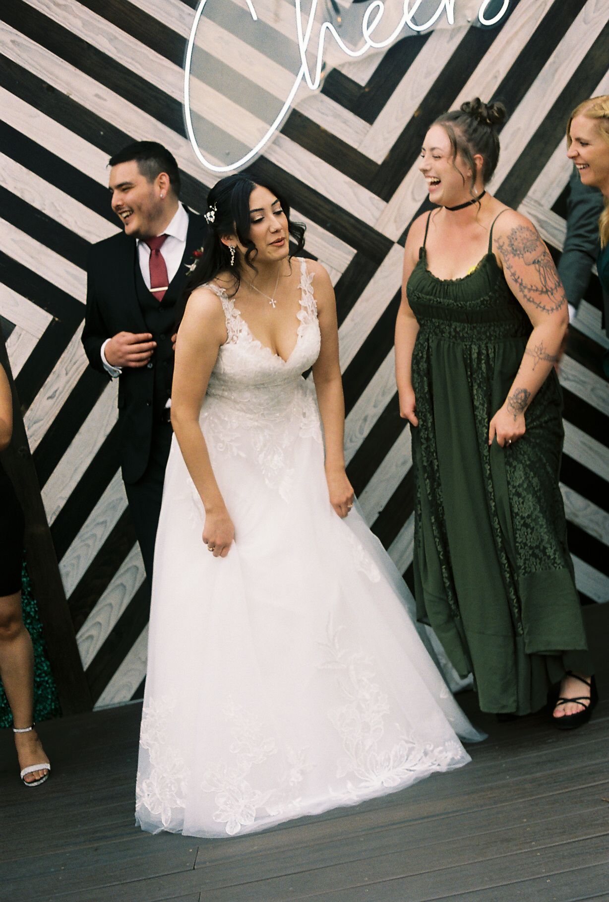 Bride in lace V-neck wedding gown laughs with groom and guest in front of striped Cheers backdrop.