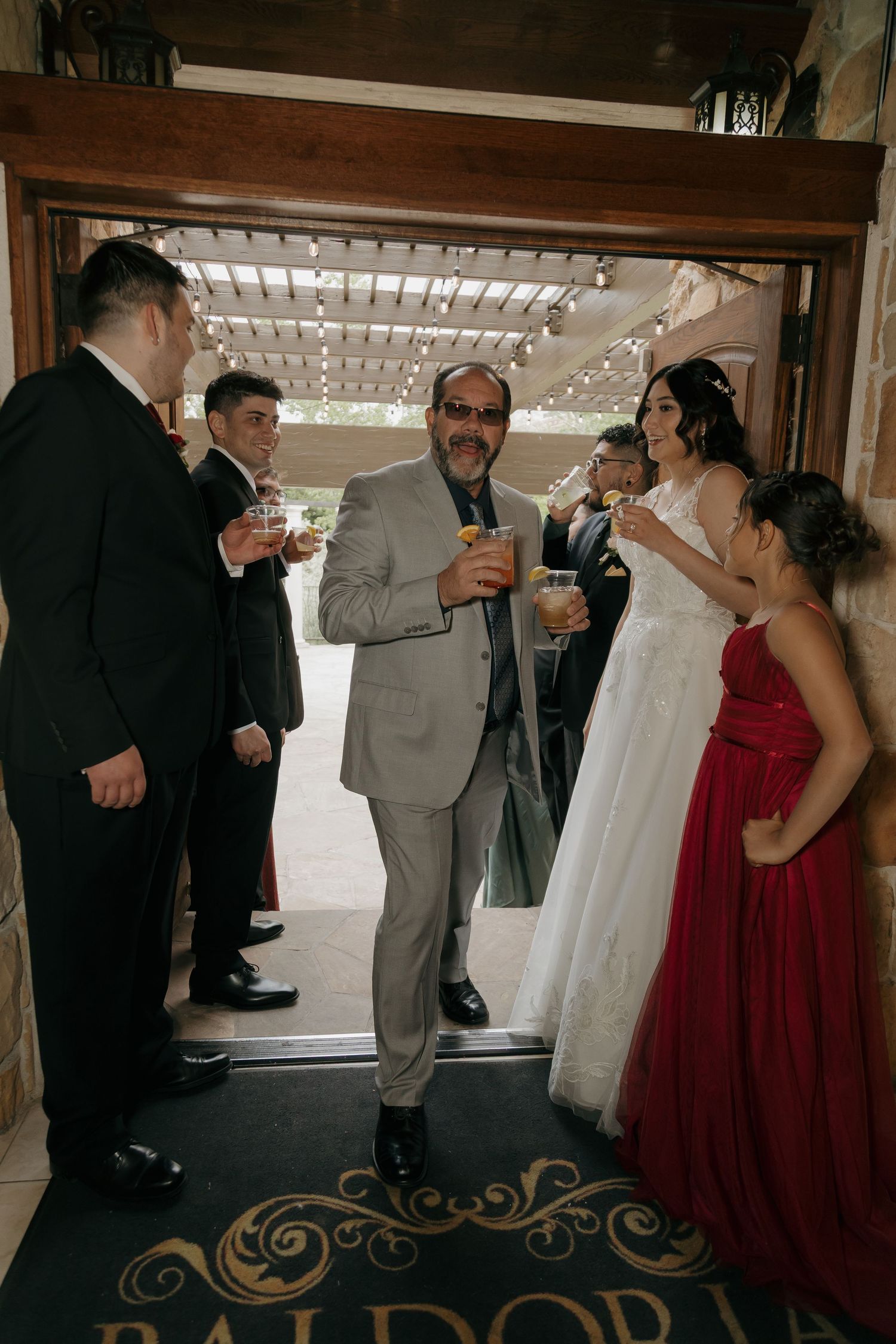 Father of the bride in gray suit holds a drink while greeting the bride and guests at venue entrance.