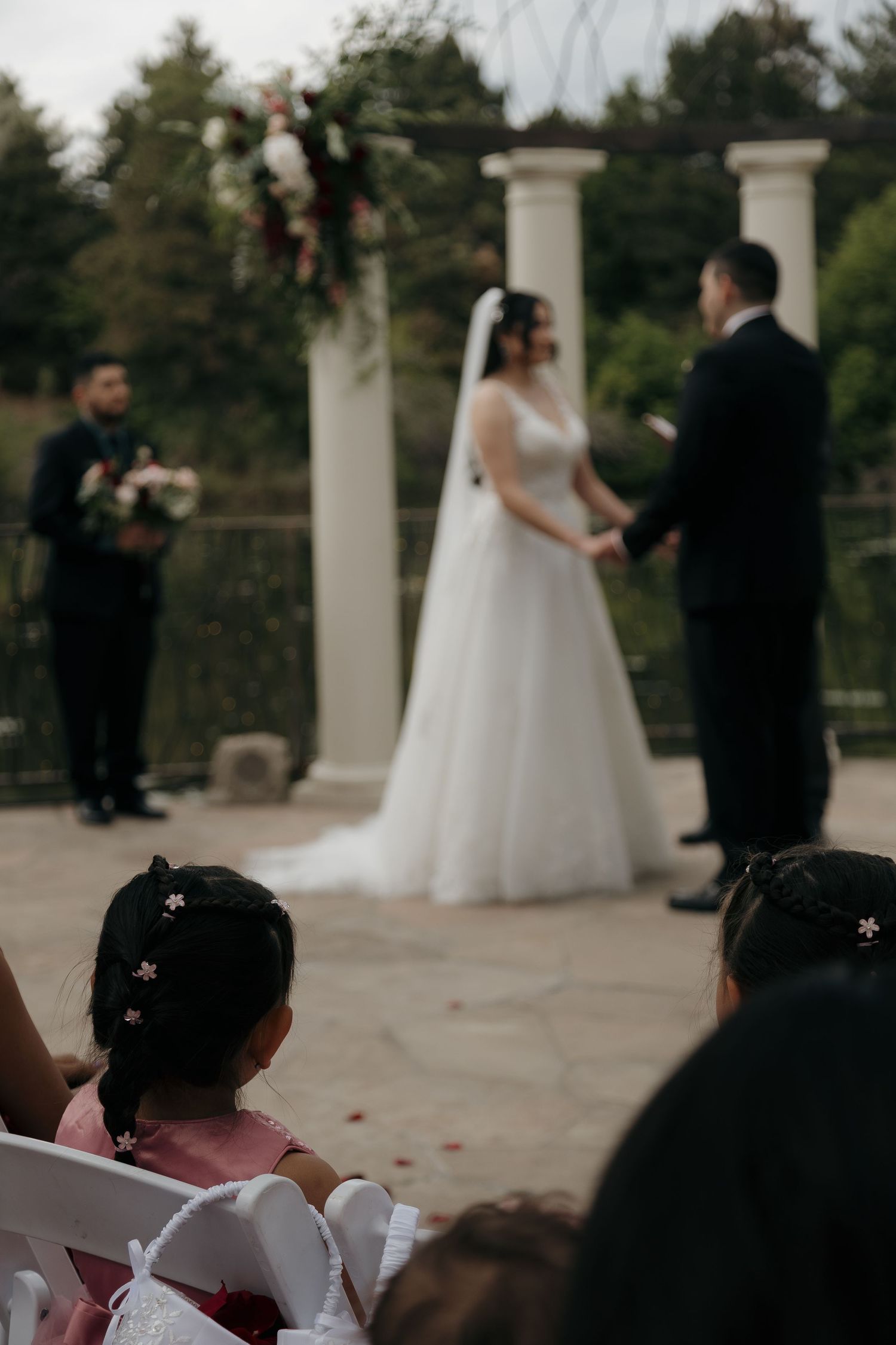 Flower girl watches bride and groom exchange vows at outdoor wedding ceremony with columns.