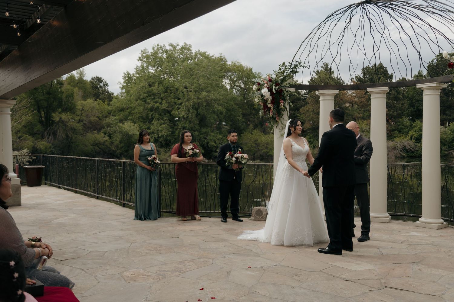 Bride and groom exchange vows at an outdoor wedding ceremony with bridesmaids and floral arch by a scenic waterfront.