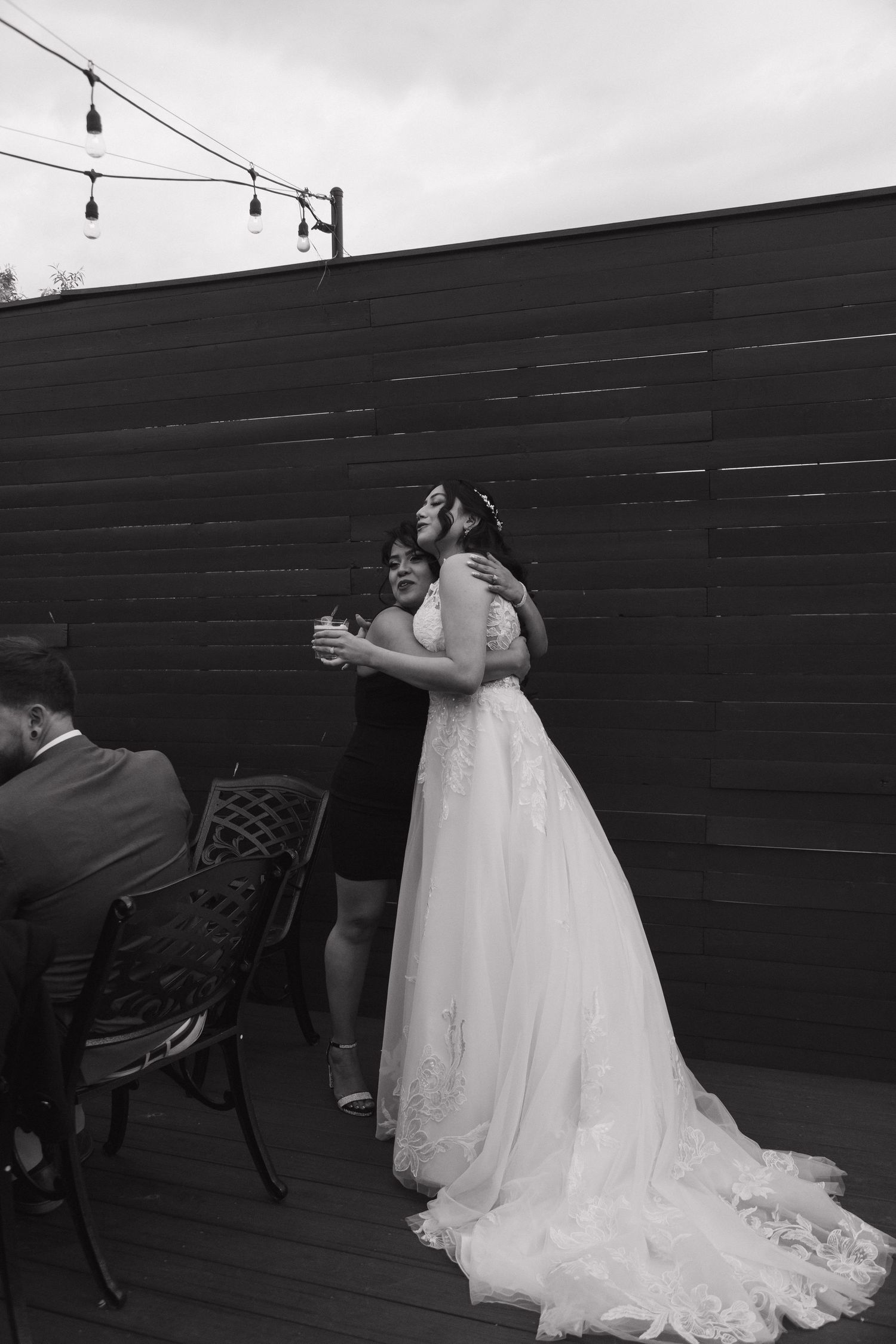 Black and white photo of bride in lace gown sharing a warm embrace with a wedding guest outdoors.