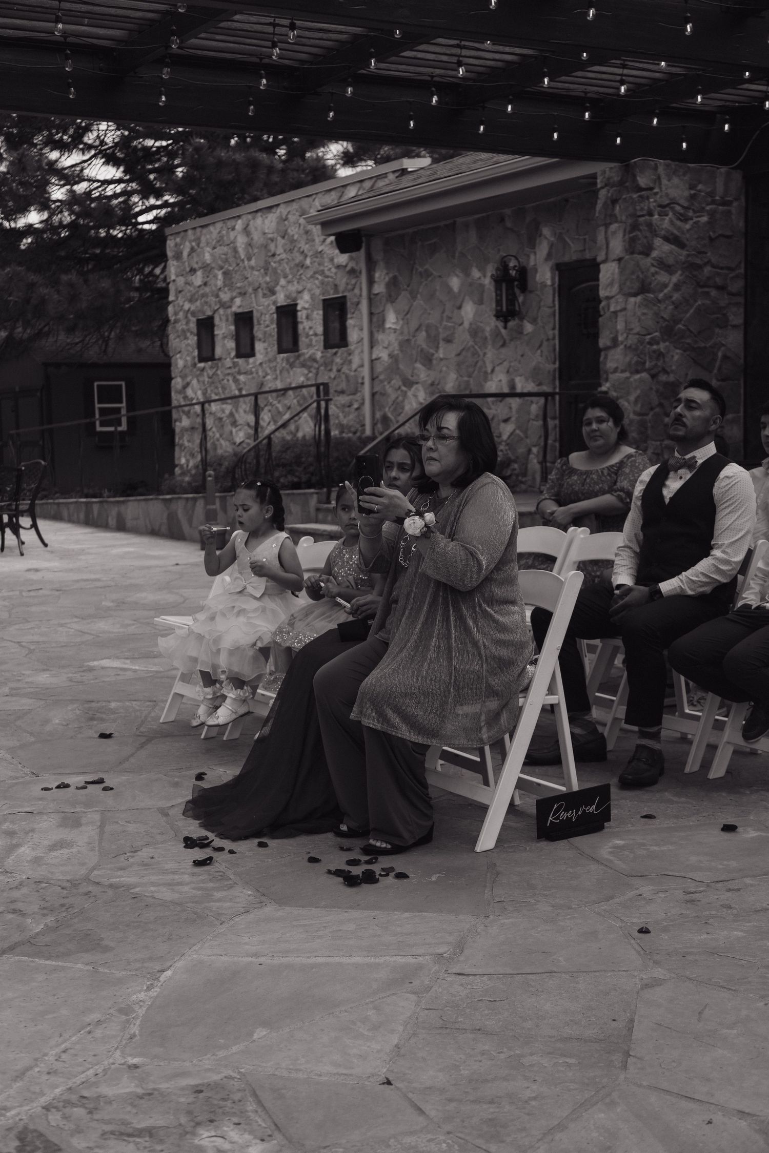 Wedding guests seated in white chairs on stone patio watch outdoor ceremony at rustic stone venue.