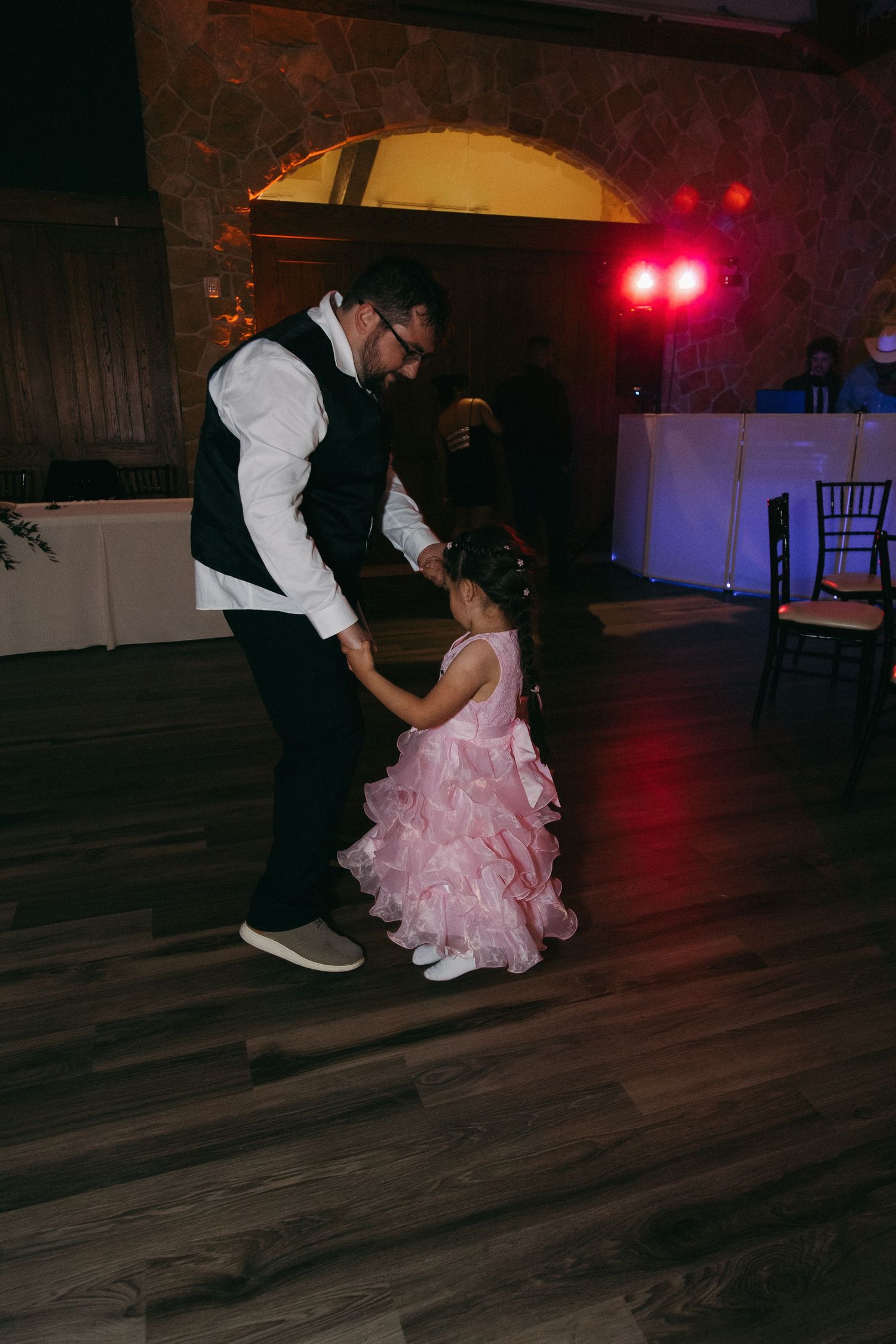 Groom in black vest dancing with a young flower girl in a pink ruffled dress on the wedding dance floor.