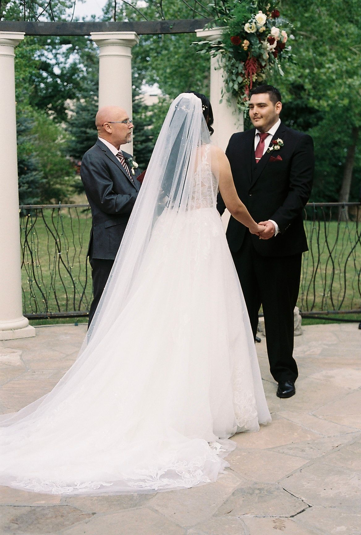 Bride in lace gown with cathedral veil stands at outdoor altar with groom and officiant.