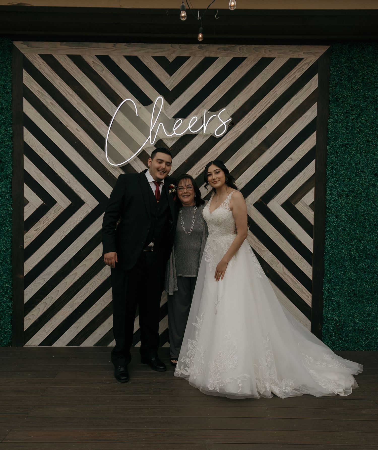 Bride and groom pose with family member in front of geometric Cheers neon sign wedding photo backdrop.