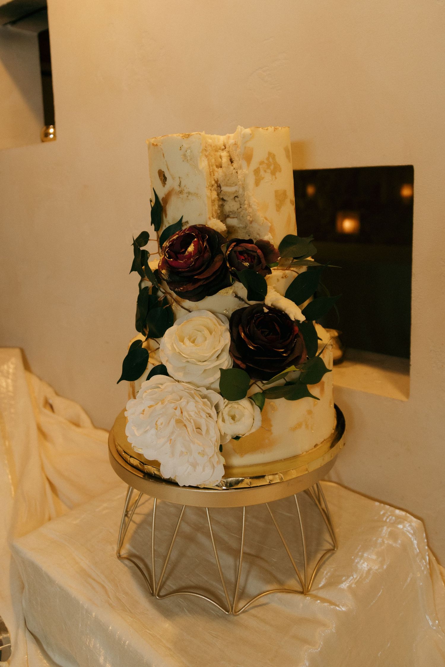 Two-tier white wedding cake with gold leaf detail and dark burgundy and white sugar roses on gold stand.