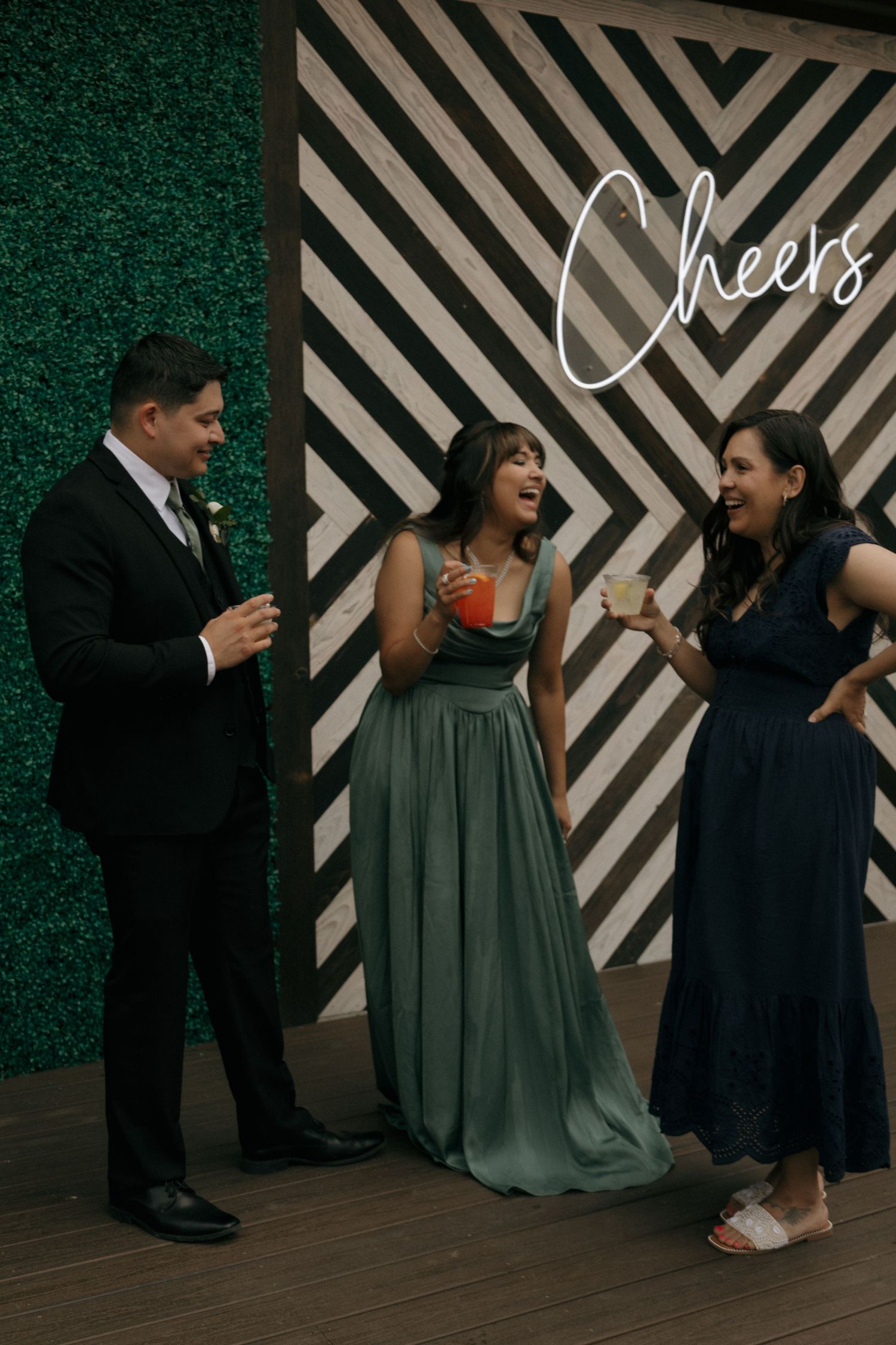Wedding guests laugh and toast drinks together in front of a geometric hedge wall with Cheers sign.