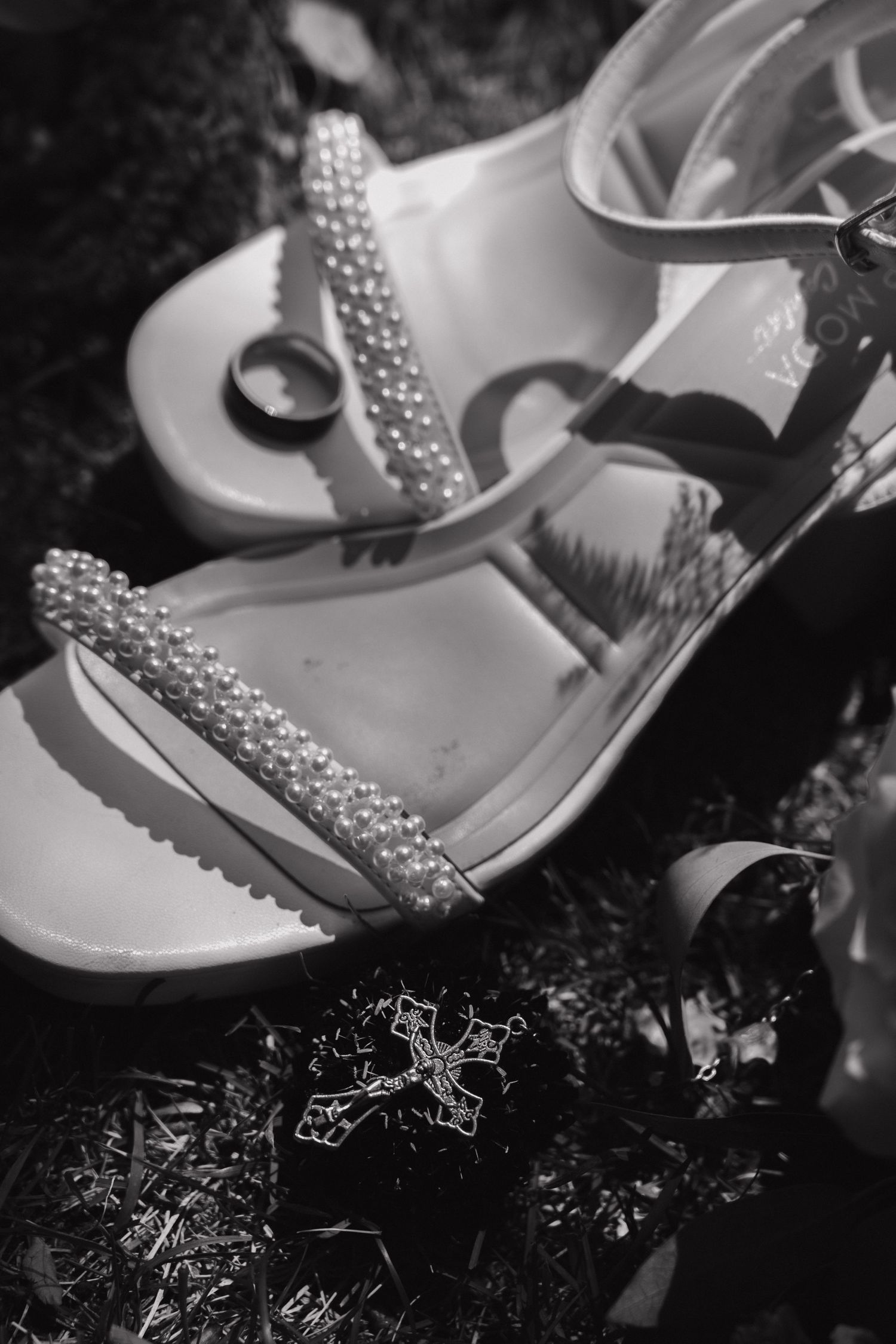 Black and white photo of pearl-embellished bridal heels with wedding ring and cross jewelry on grass.