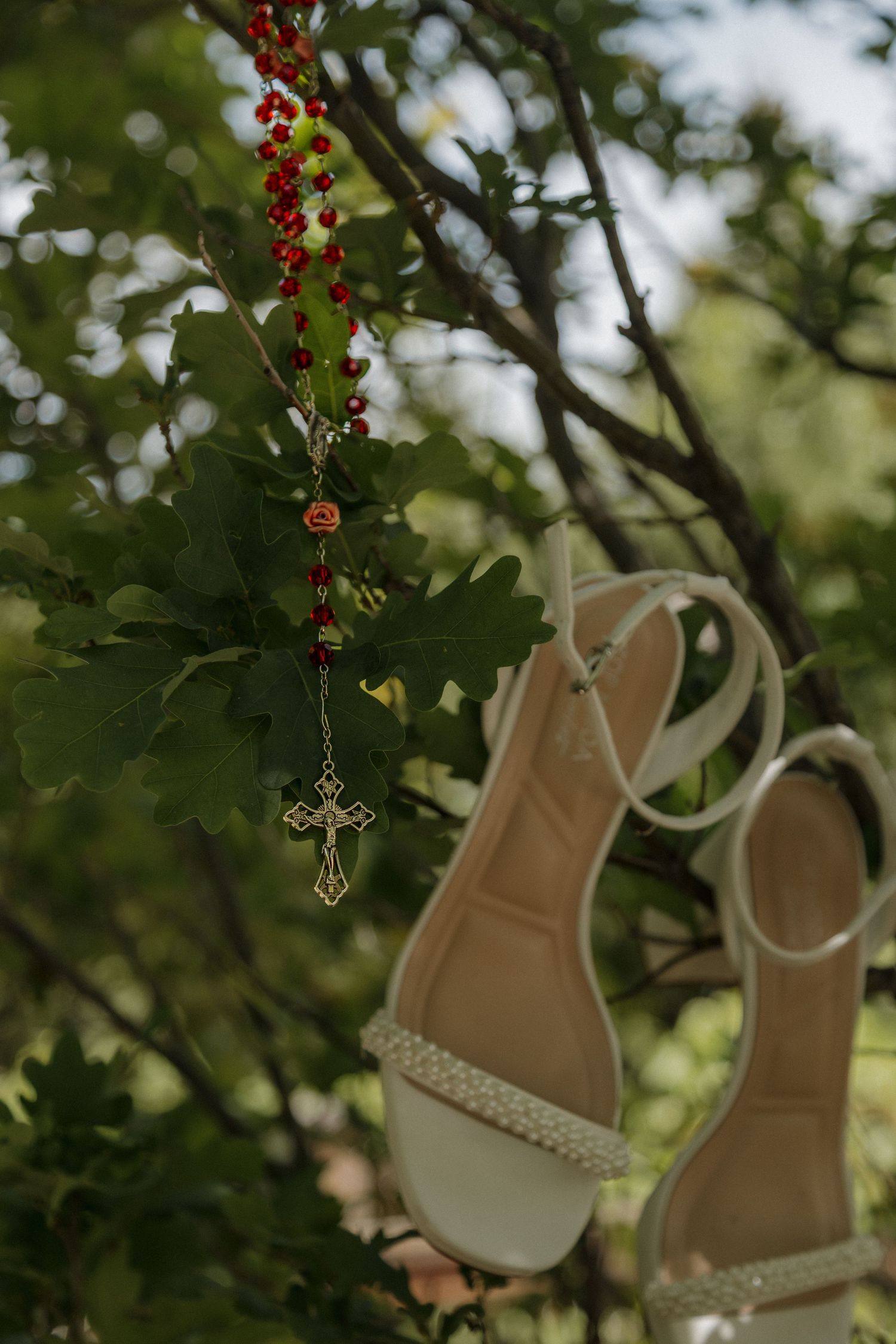 White bridal heels and red rosary beads with gold cross hanging from a green tree branch outdoors.