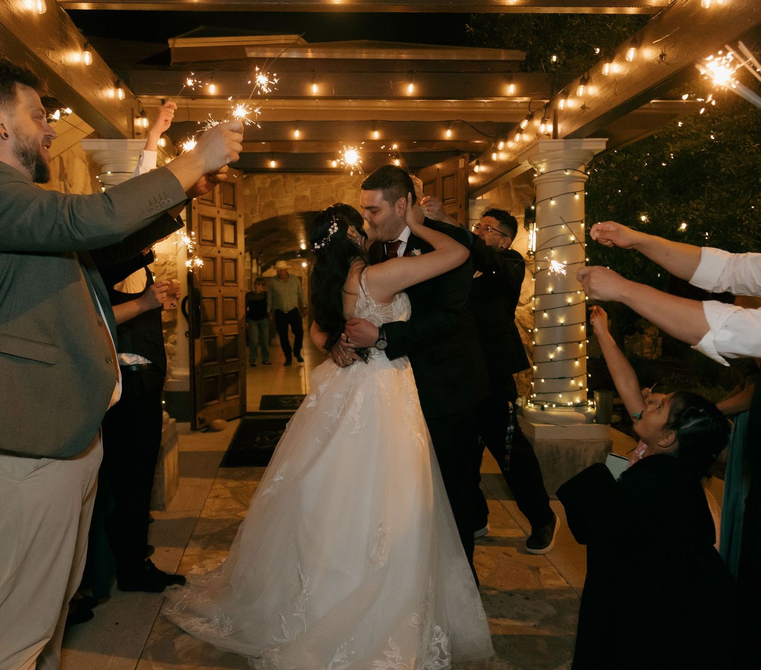 Bride and groom embrace during a dazzling sparkler send-off under warm string lights at night.