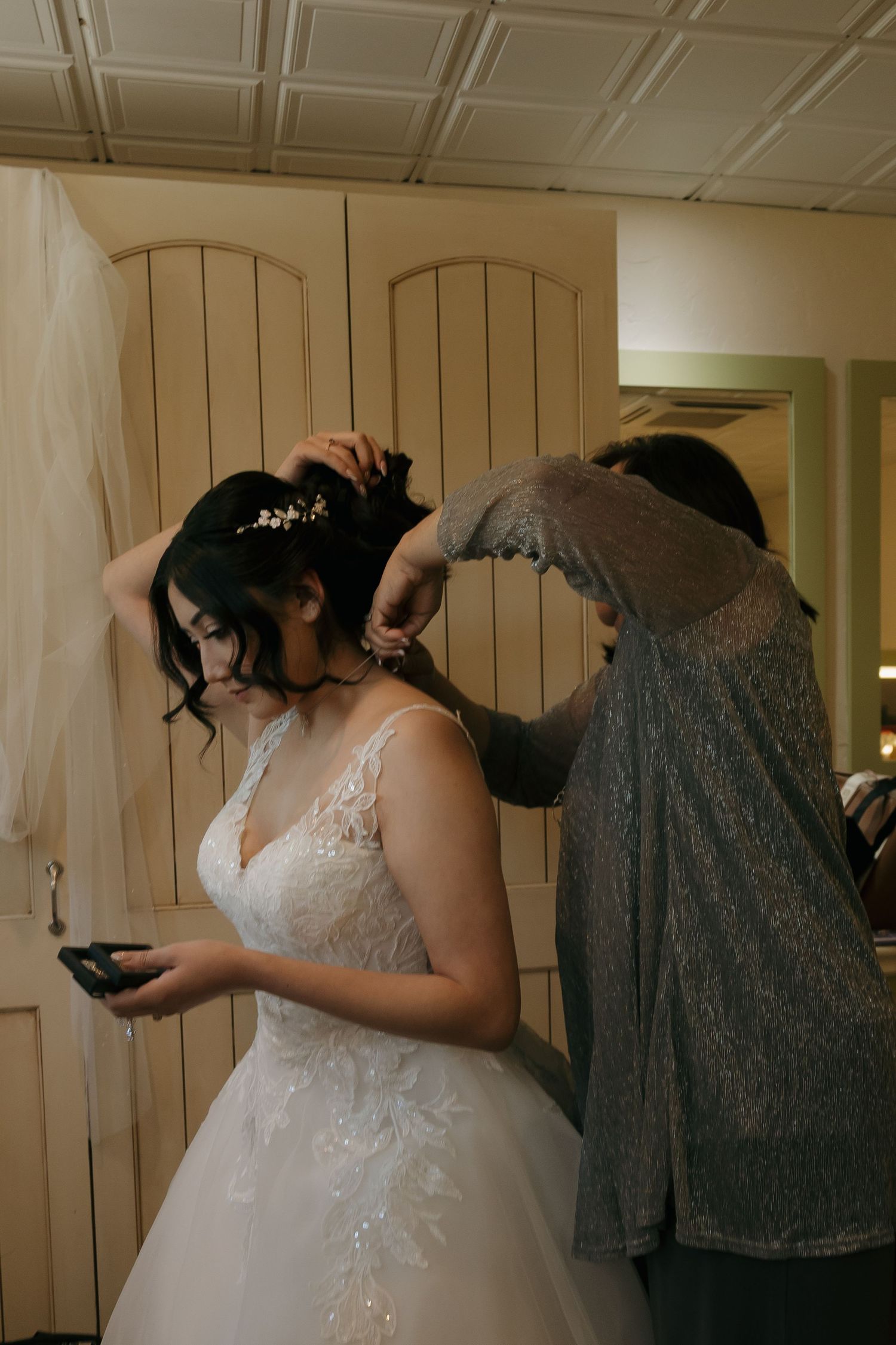 Bride in lace wedding gown getting hair styled by assistant before ceremony.