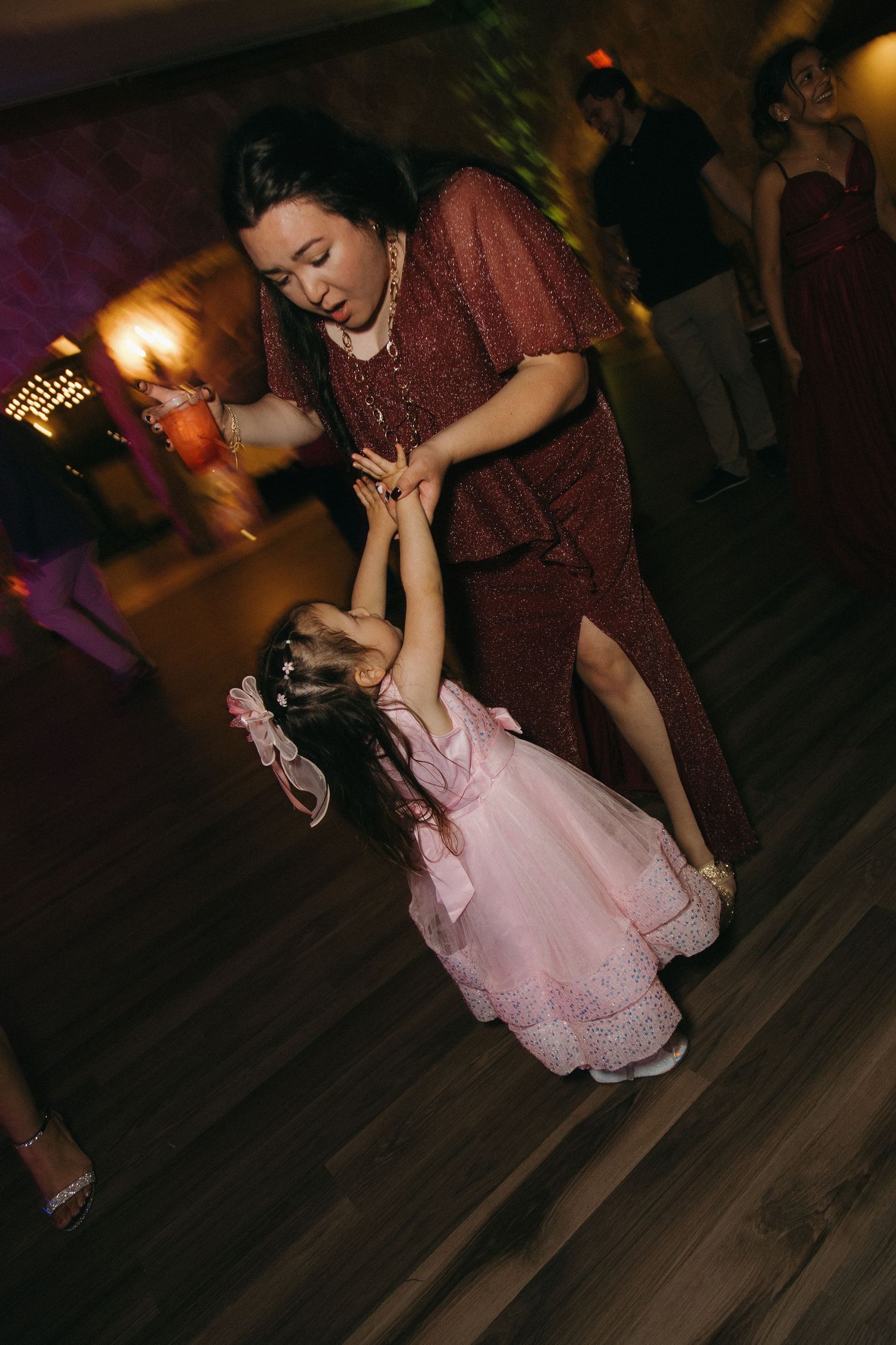 Woman in burgundy dress dances joyfully with young girl in pink at wedding reception.