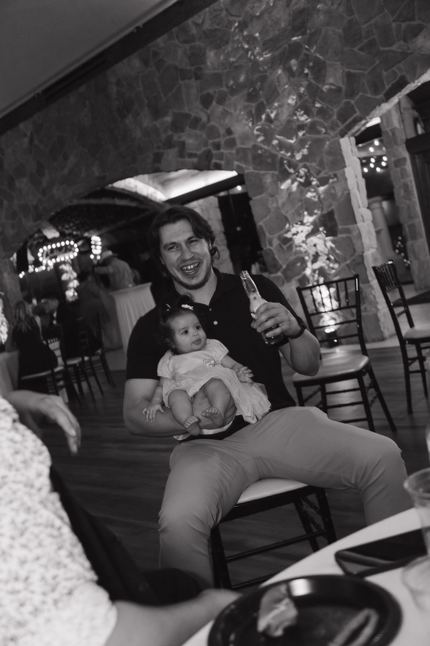 Happy father holding baby at wedding reception in black and white photo at rustic stone venue.